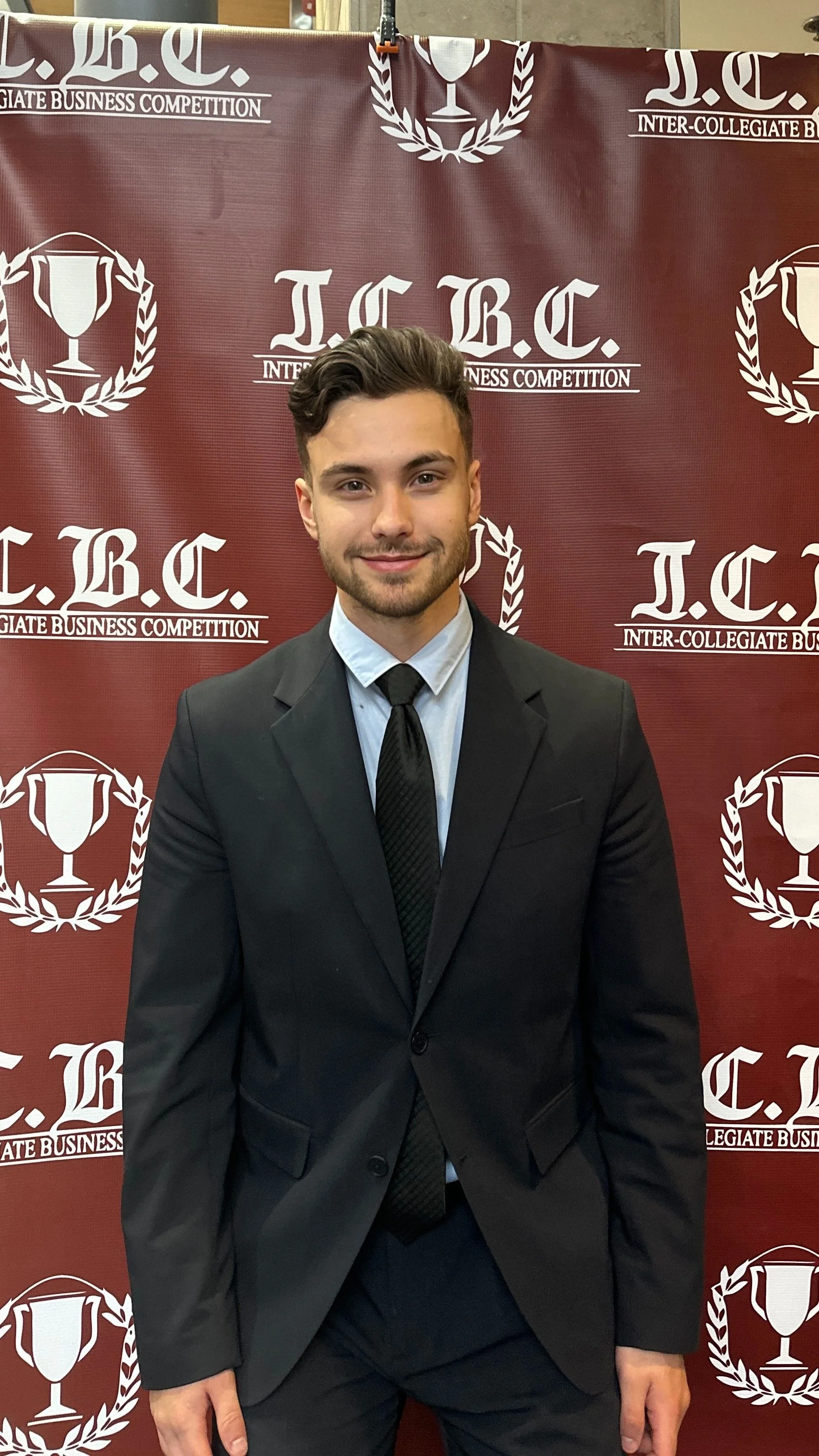 A young man in a suit standing in front of a maroon backdrop with white logos and text that read 'I.C. Inter-Collegiate Business Competition' and images of trophies.
