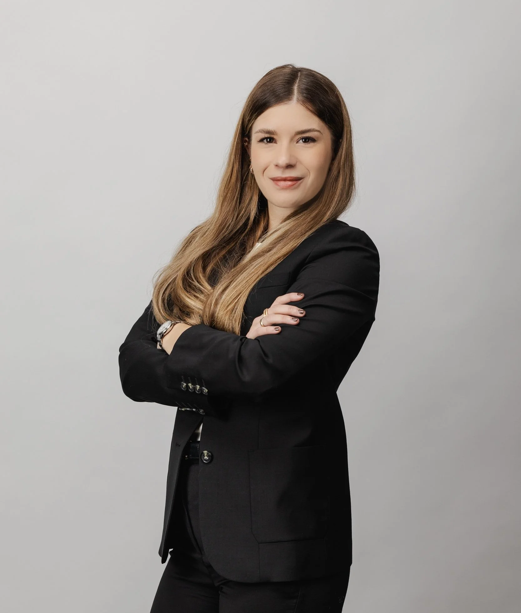 Femme habillée en noir, cheveux longs, souriant, croisant les bras, fond gris clair.