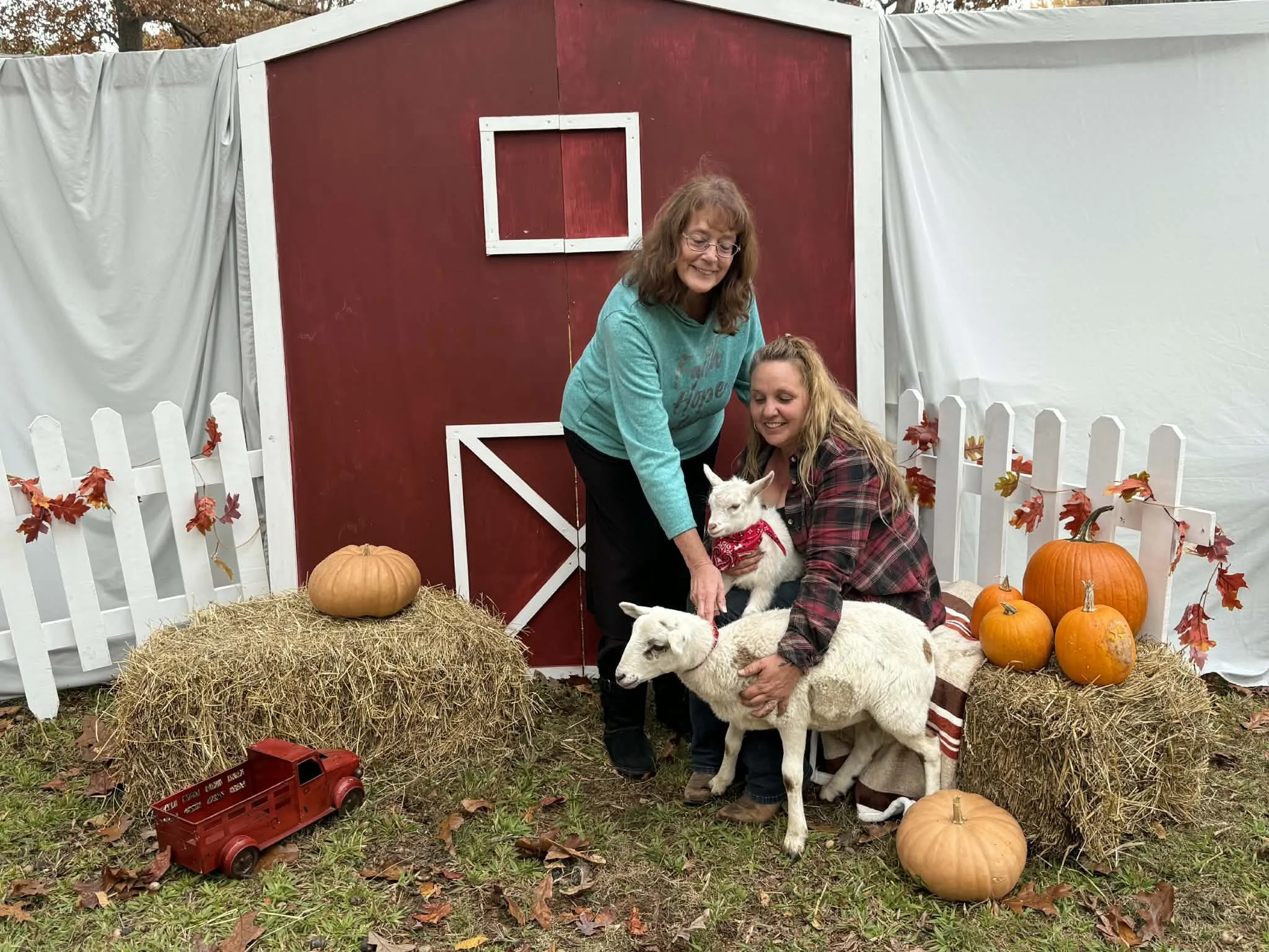Two women with two goats, pumpkins, hay bales, a red barn backdrop, and a white picket fence, celebrating fall or Halloween outdoors.