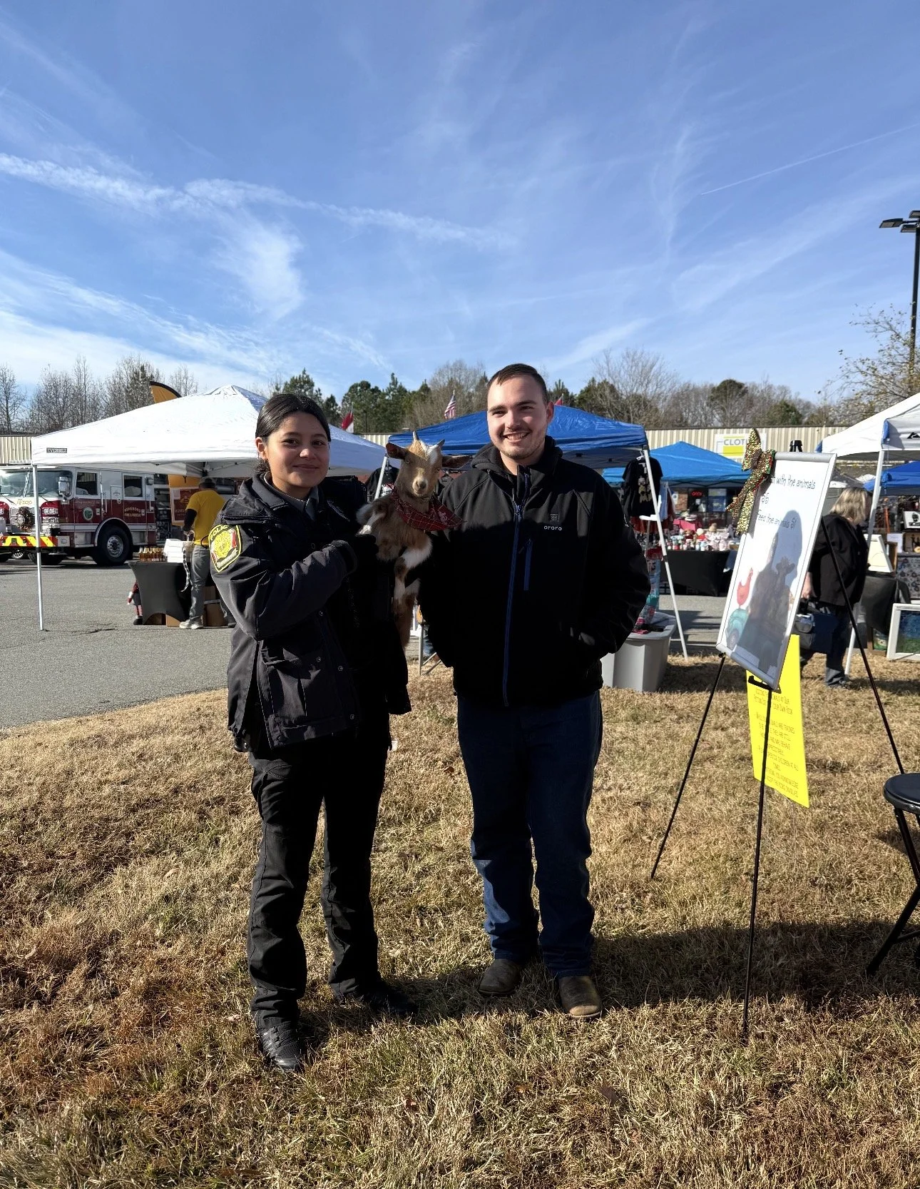 Two people, one in a police uniform and the other in a black jacket, stand outdoors at a market or fair. The person in uniform holds a small brown and white goat. Descriptive signs and tents are visible in the background.