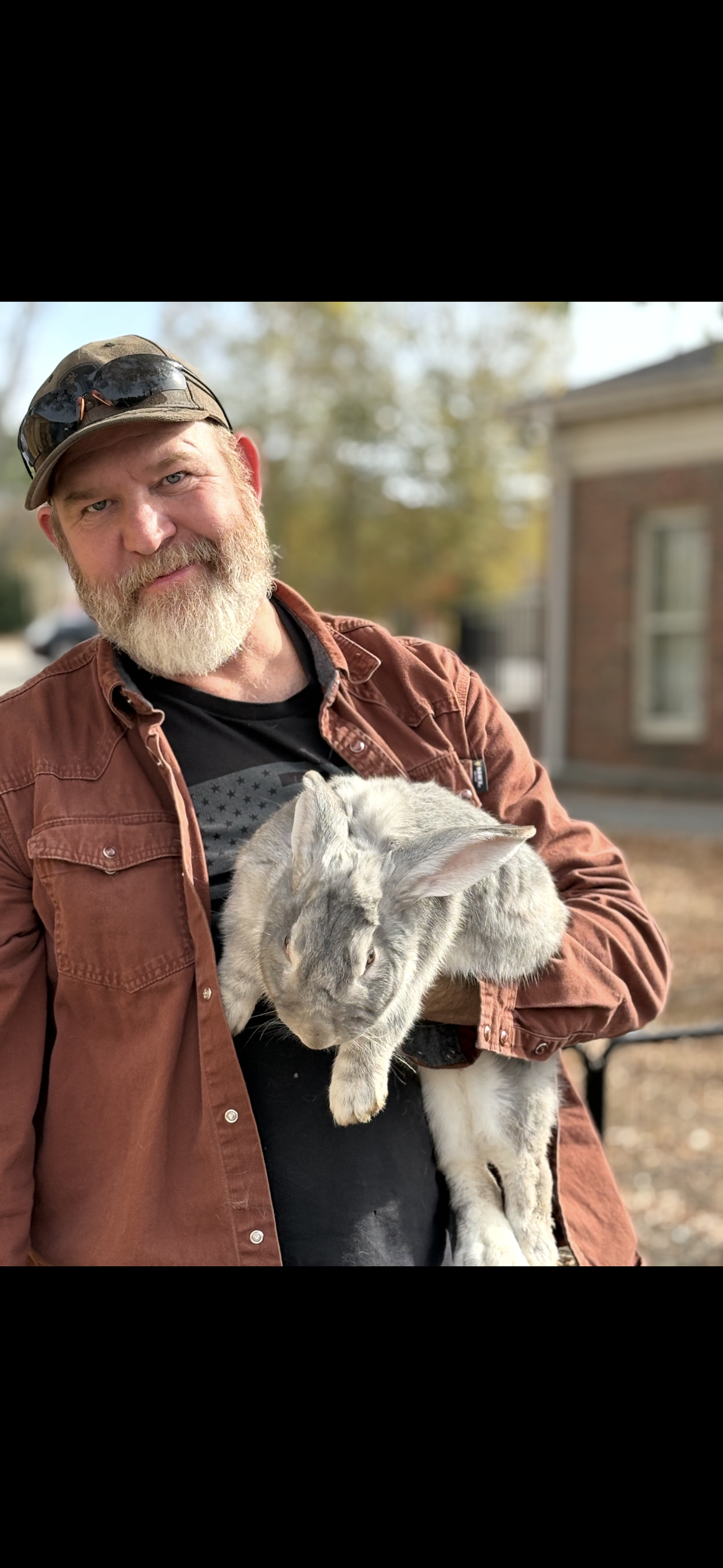 A man with a beard wearing a cap, sunglasses, and a brown jacket, holding a gray rabbit outdoors.