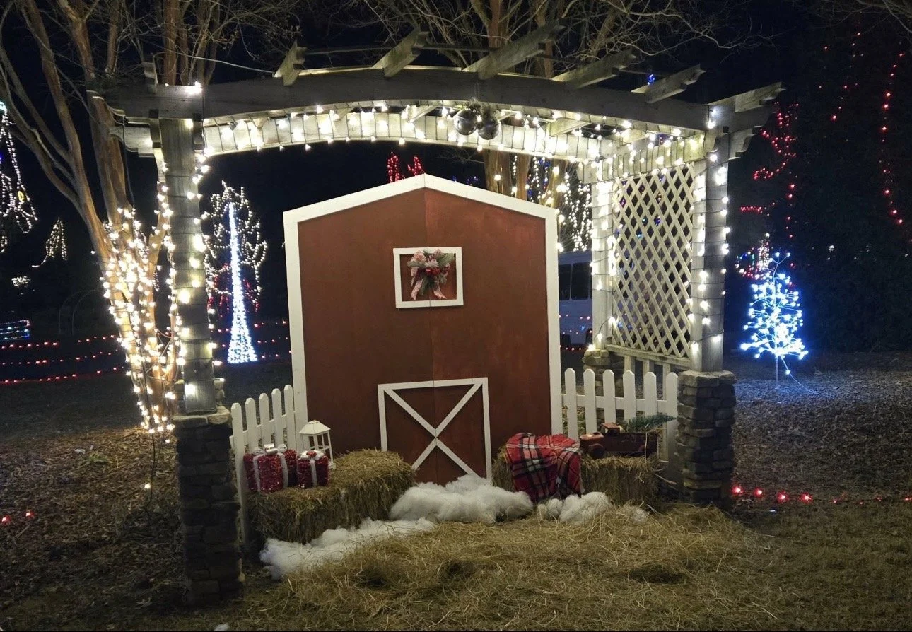 Outdoor Christmas nativity scene with a decorated stable, hay, wrapped presents, and illuminated trees and lights at night.