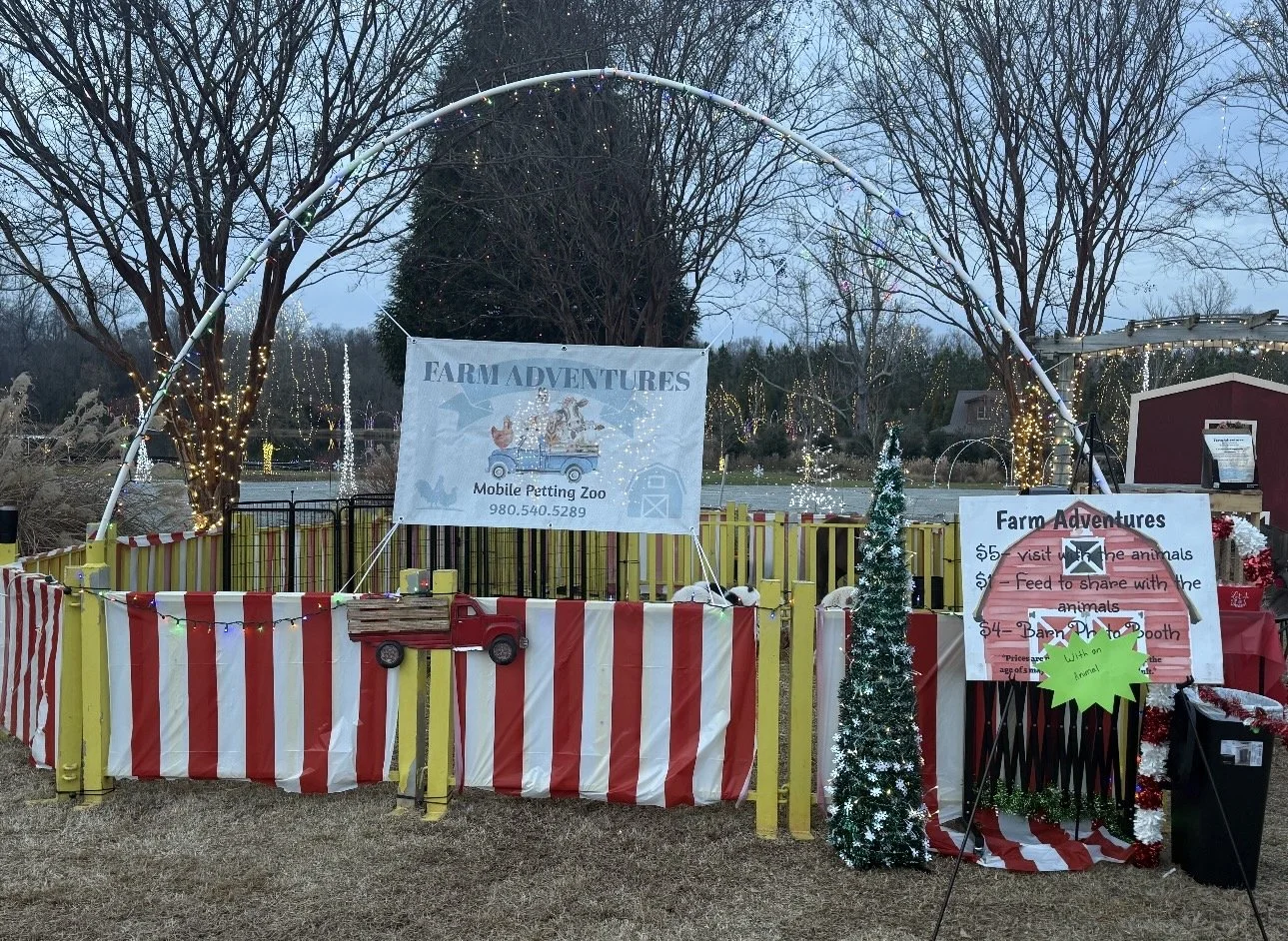 Outdoor farm-themed attraction with a yellow and red striped fence, decorated with Christmas lights and small trees, advertising a mobile petting zoo. Signs indicate entry fee of $5, with additional activities such as feeding animals and a photo boot