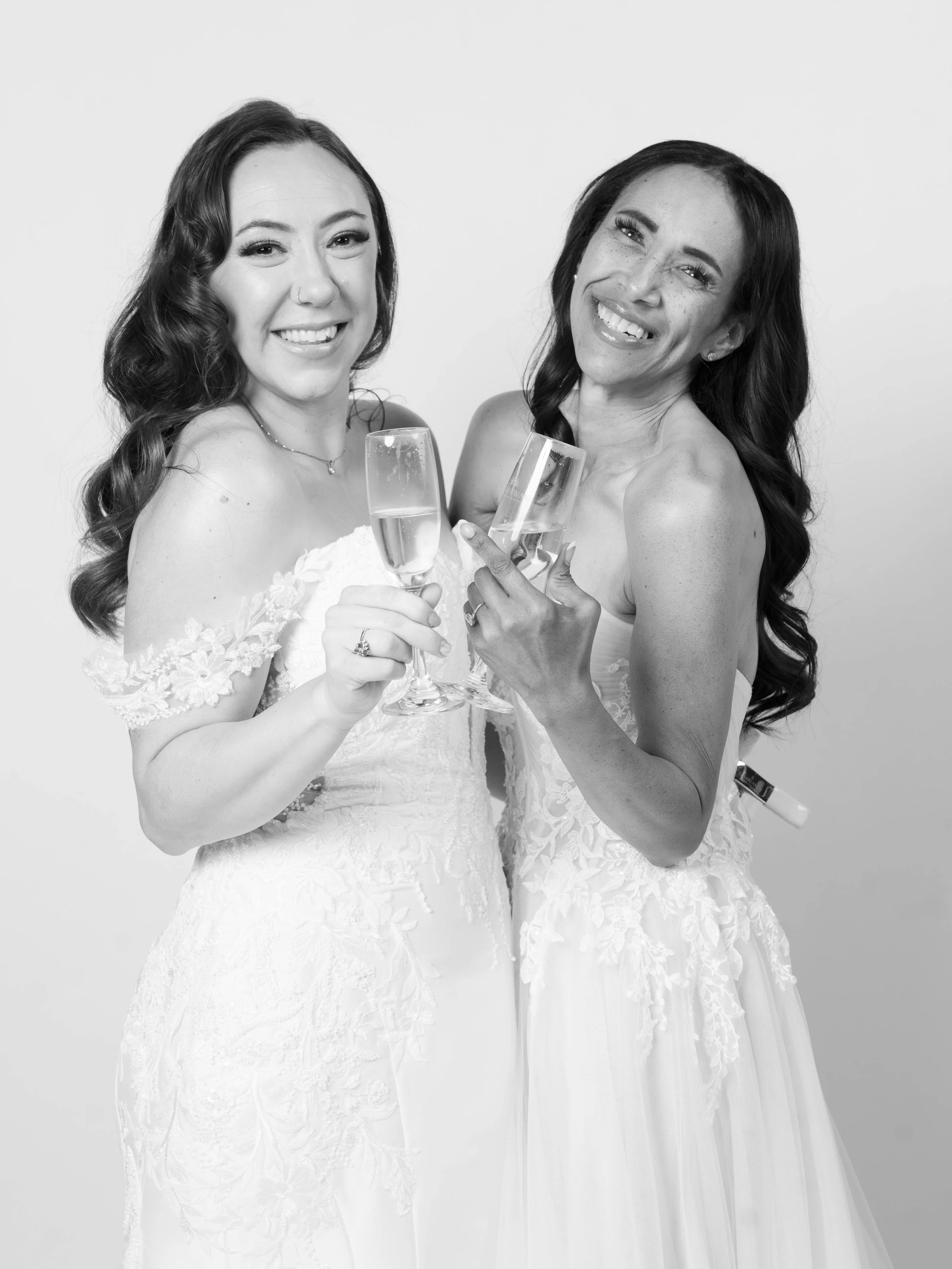 Two women in wedding dresses holding glasses of champagne and smiling at the camera.