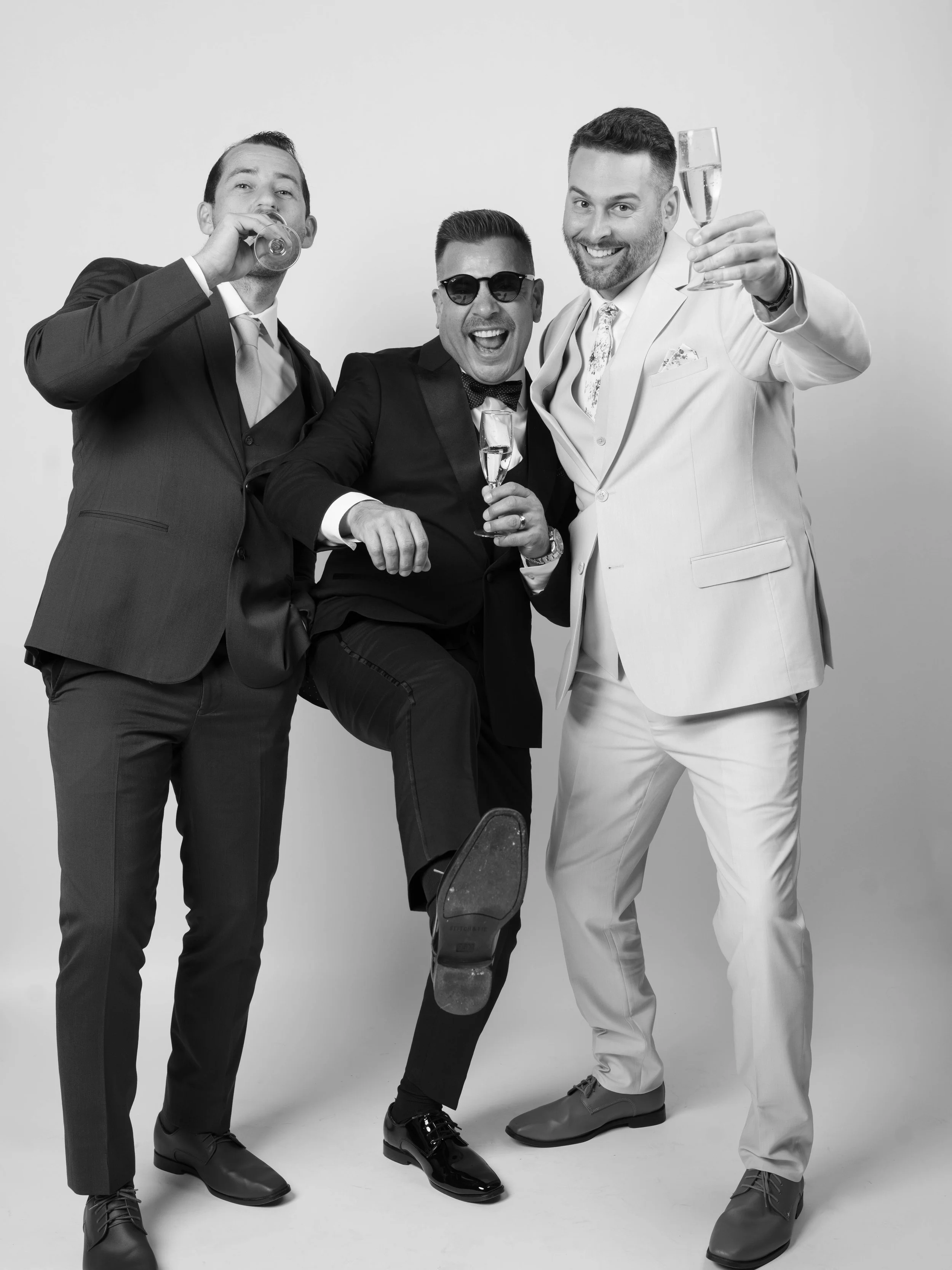 Black and white photo of three men in suits celebrating, holding champagne glasses, with one man in sunglasses and the middle man kicking his leg up.