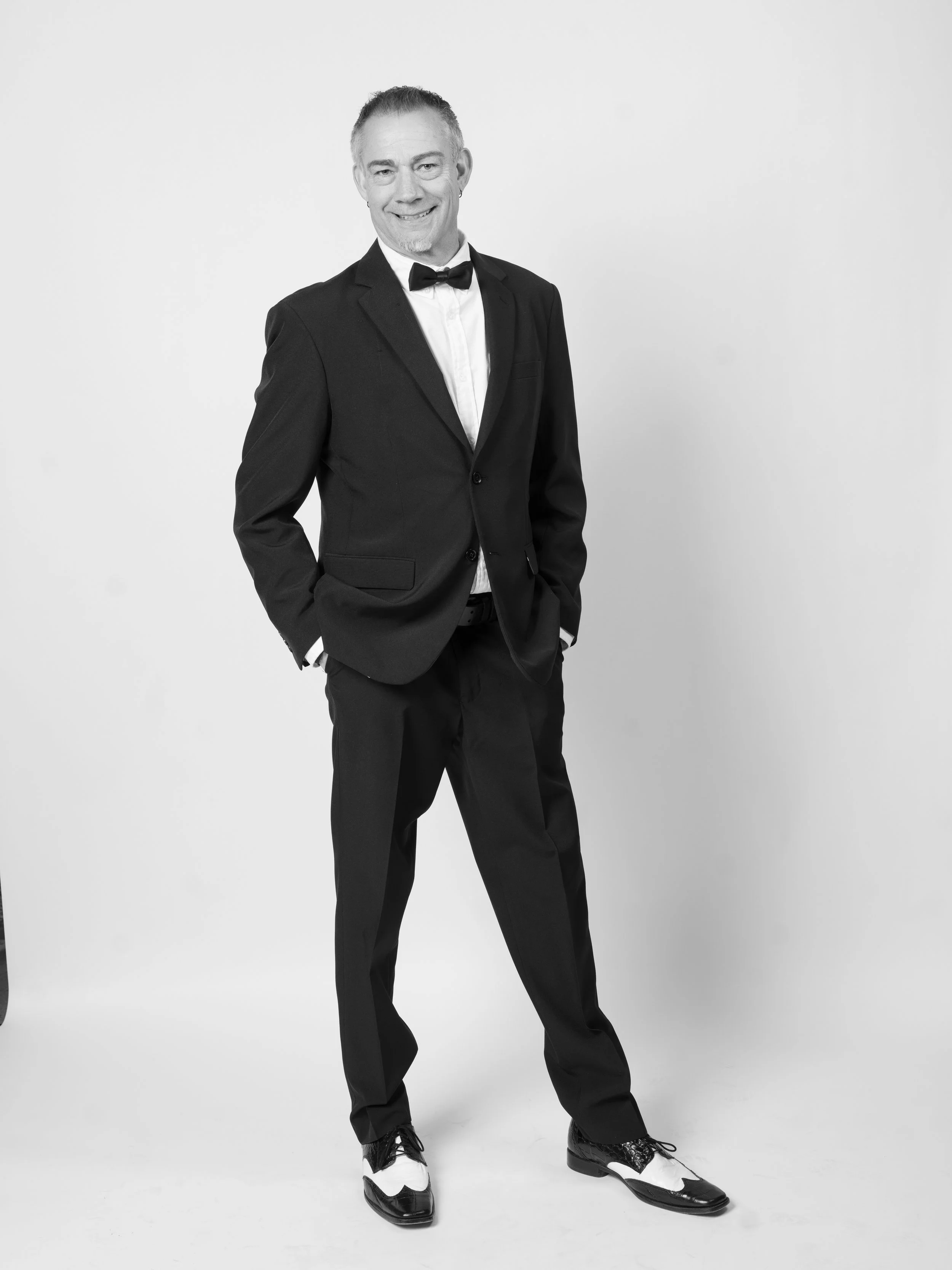 Man in tuxedo with bow tie and two-tone tuxedo shoes standing against a plain background.