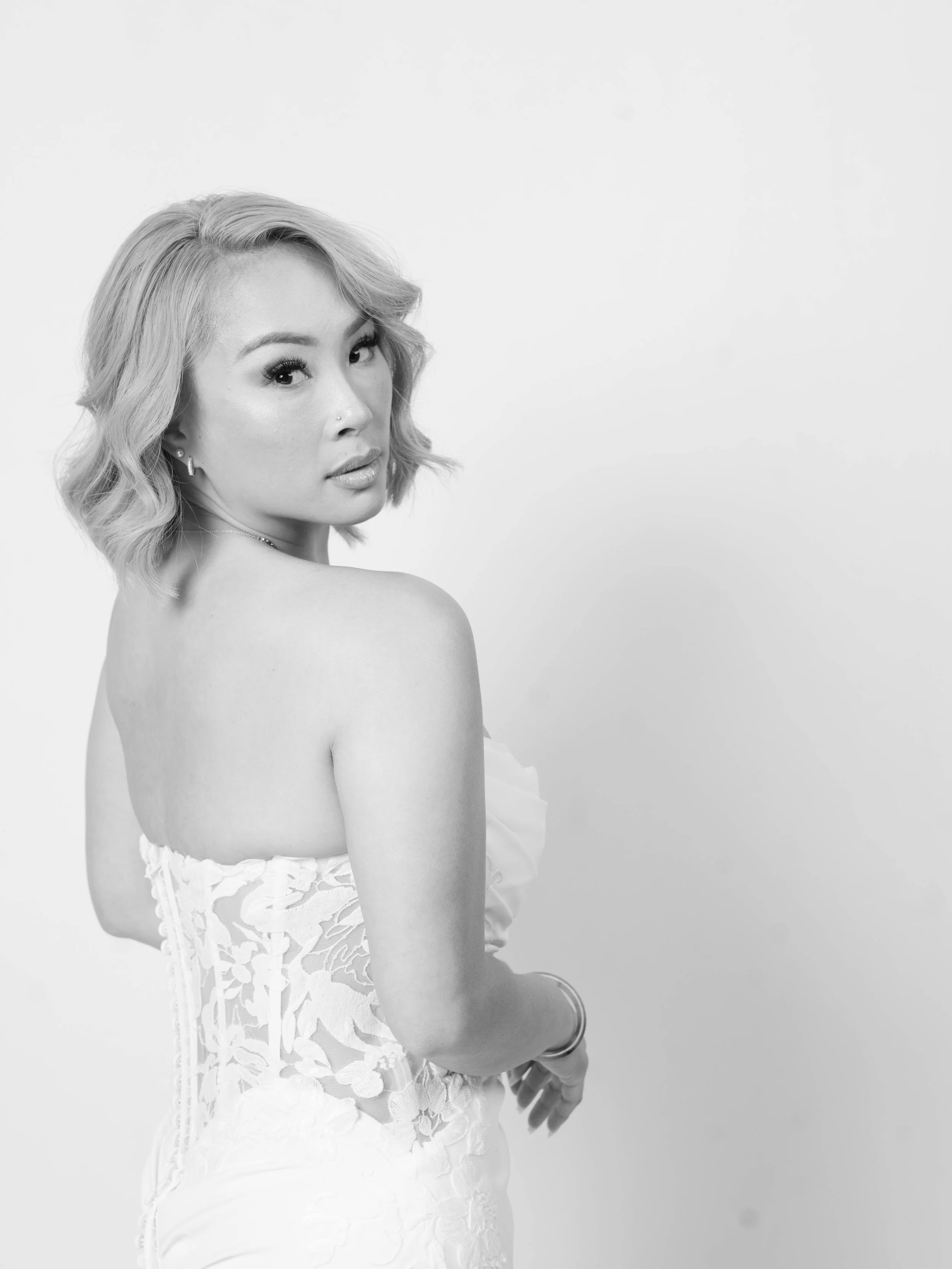 Black and white portrait of a young woman with short wavy hair, looking over her shoulder at the camera, wearing a strapless lace dress.