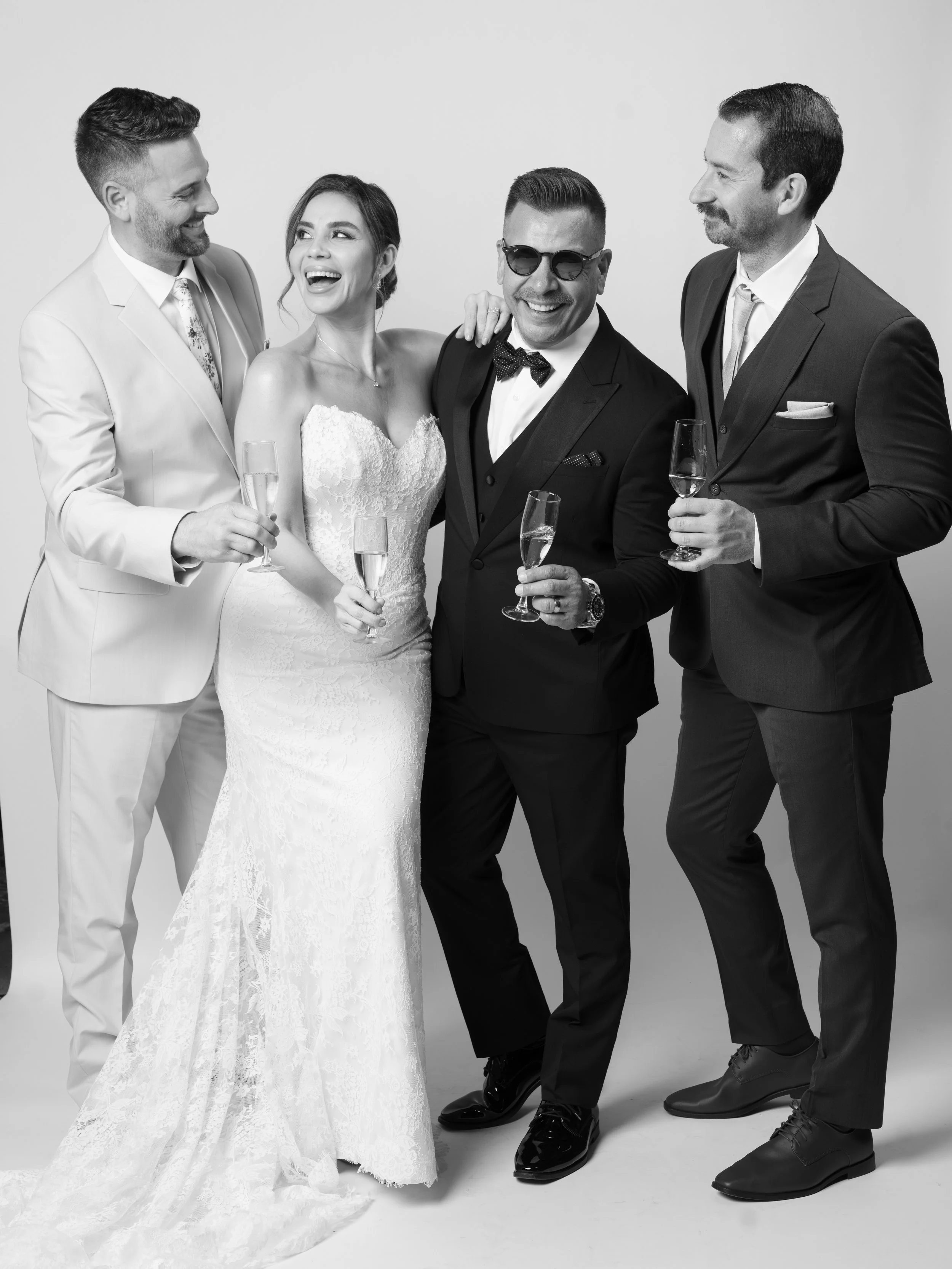 Four people in formal attire celebrating, with three men in suits and a woman in a wedding dress, holding champagne glasses and smiling.