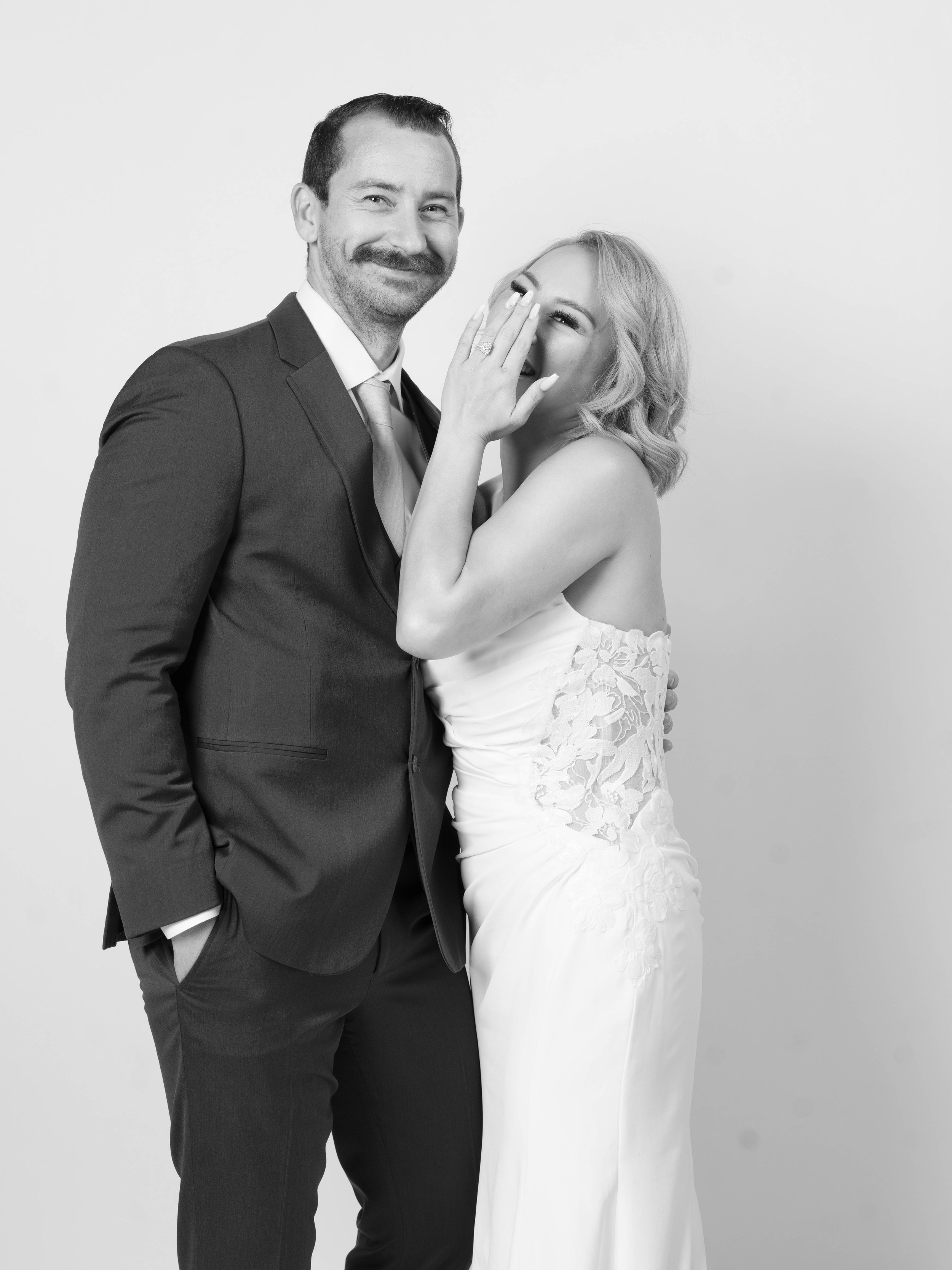 A black and white photo of a bride and groom. The bride is laughing and covering her mouth with her hand, and the groom is smiling. The bride is wearing a strapless wedding dress, and the groom is in a suit and tie.