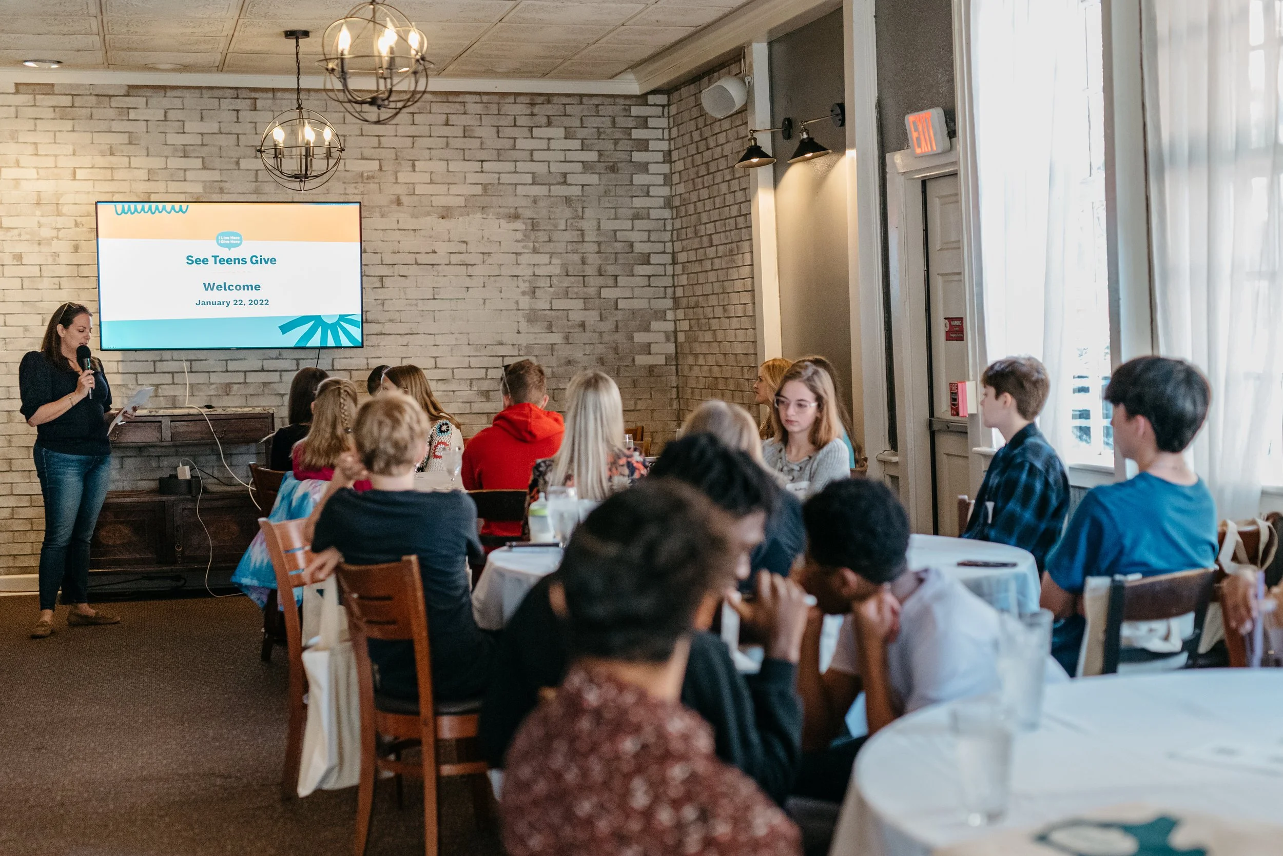 A group of teenagers sitting at round tables in a brightly lit room, watching a presentation for the See Teens Give program.