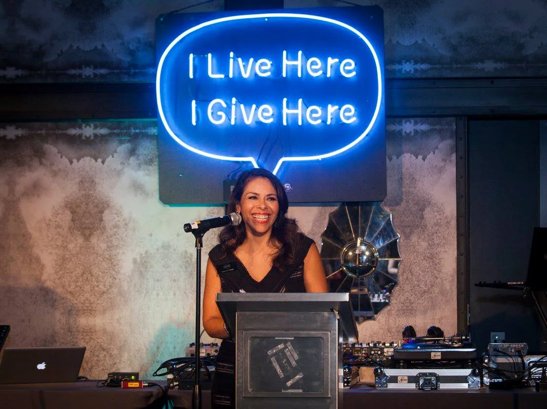 A woman speaking at a podium during an event, with a blue neon "I Live Here I Give Here" sign glowing on the wall behind her.