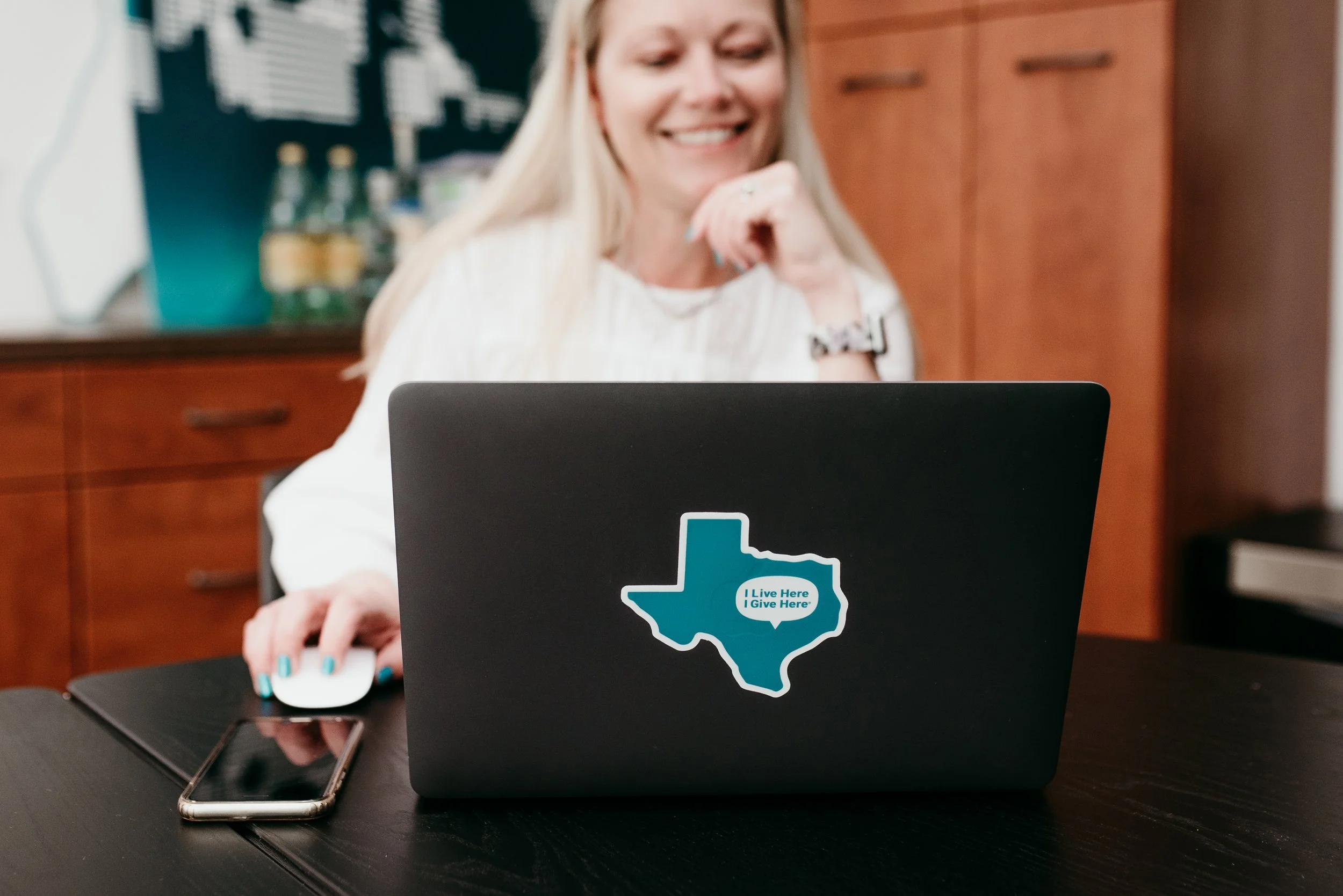 A person smiling while working on a laptop that features a teal Texas-shaped I Live Here I Give Here sticker with a heart icon.