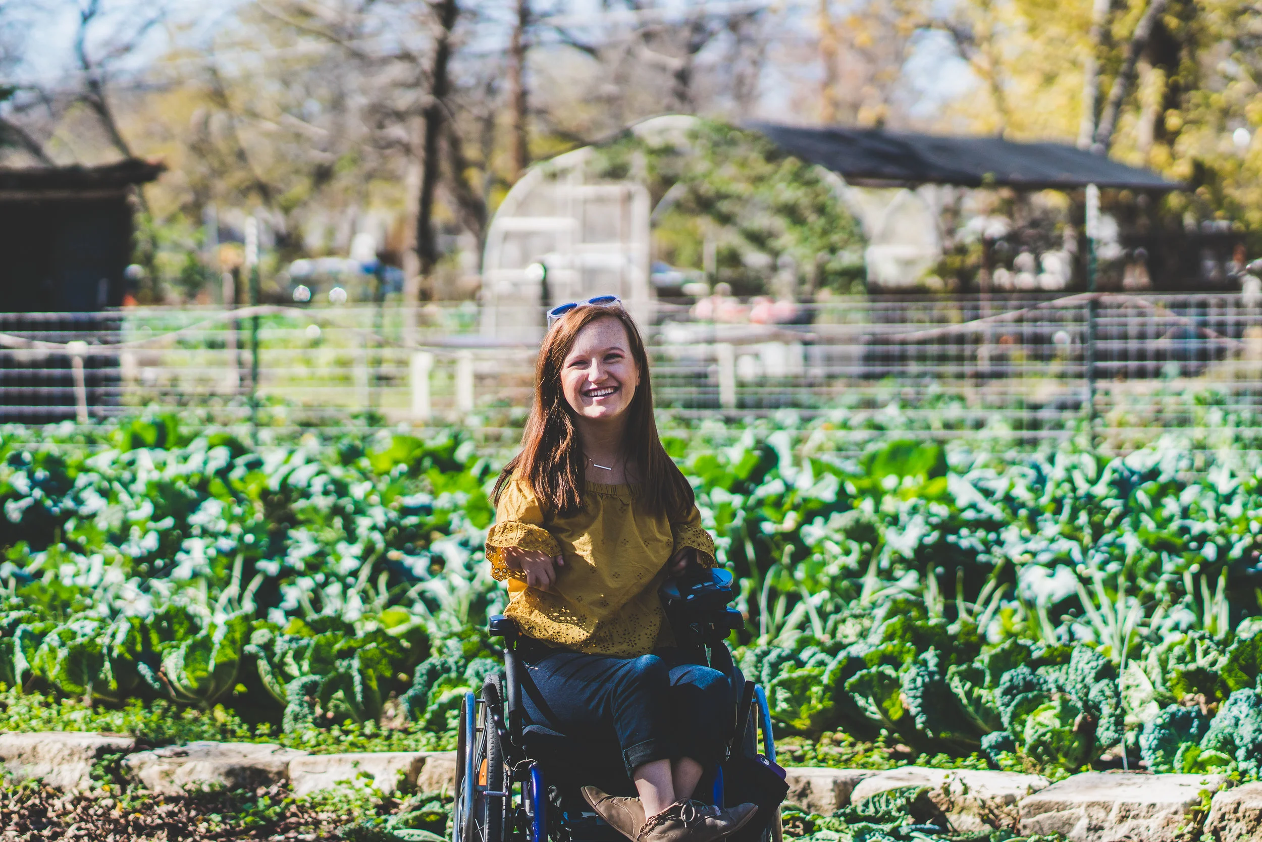 A smiling woman using a wheelchair posing in front of a lush community garden.