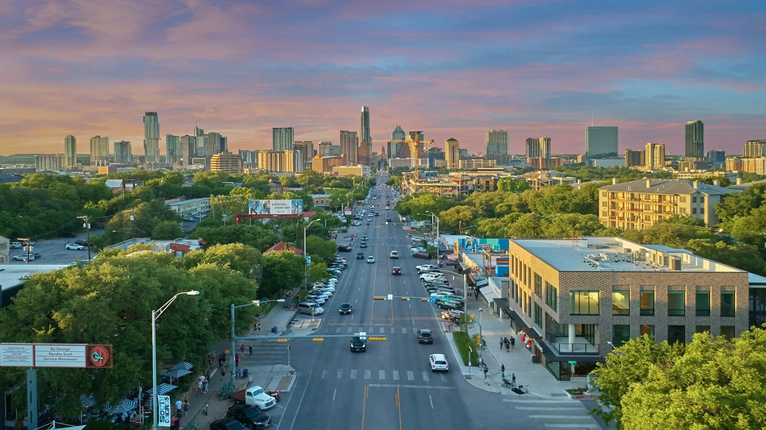 A circular aerial view of a long city street in Austin leading toward the downtown skyline at sunset.