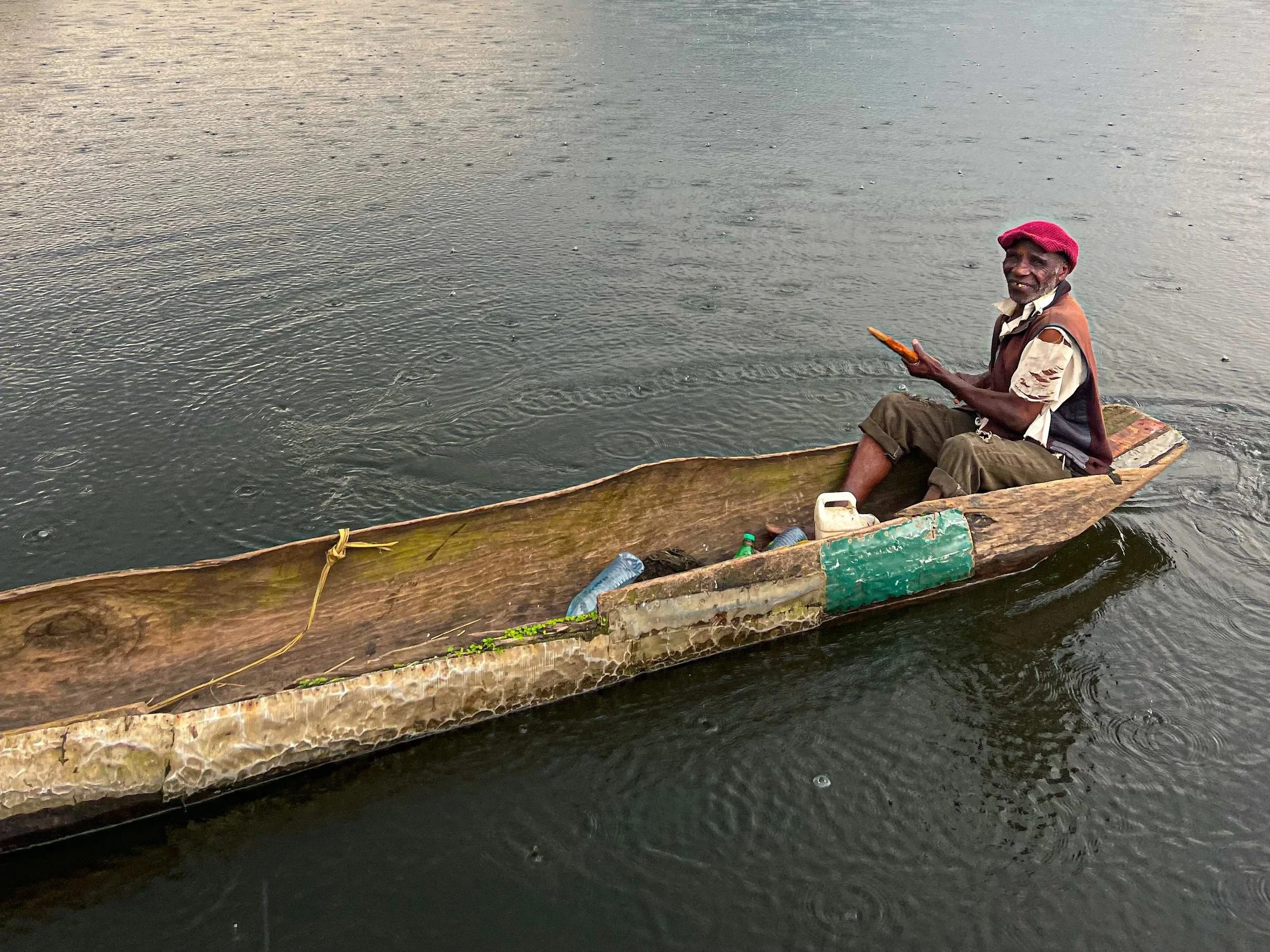 An older man sitting in a small wooden boat on the water, wearing a red hat and a brown vest.