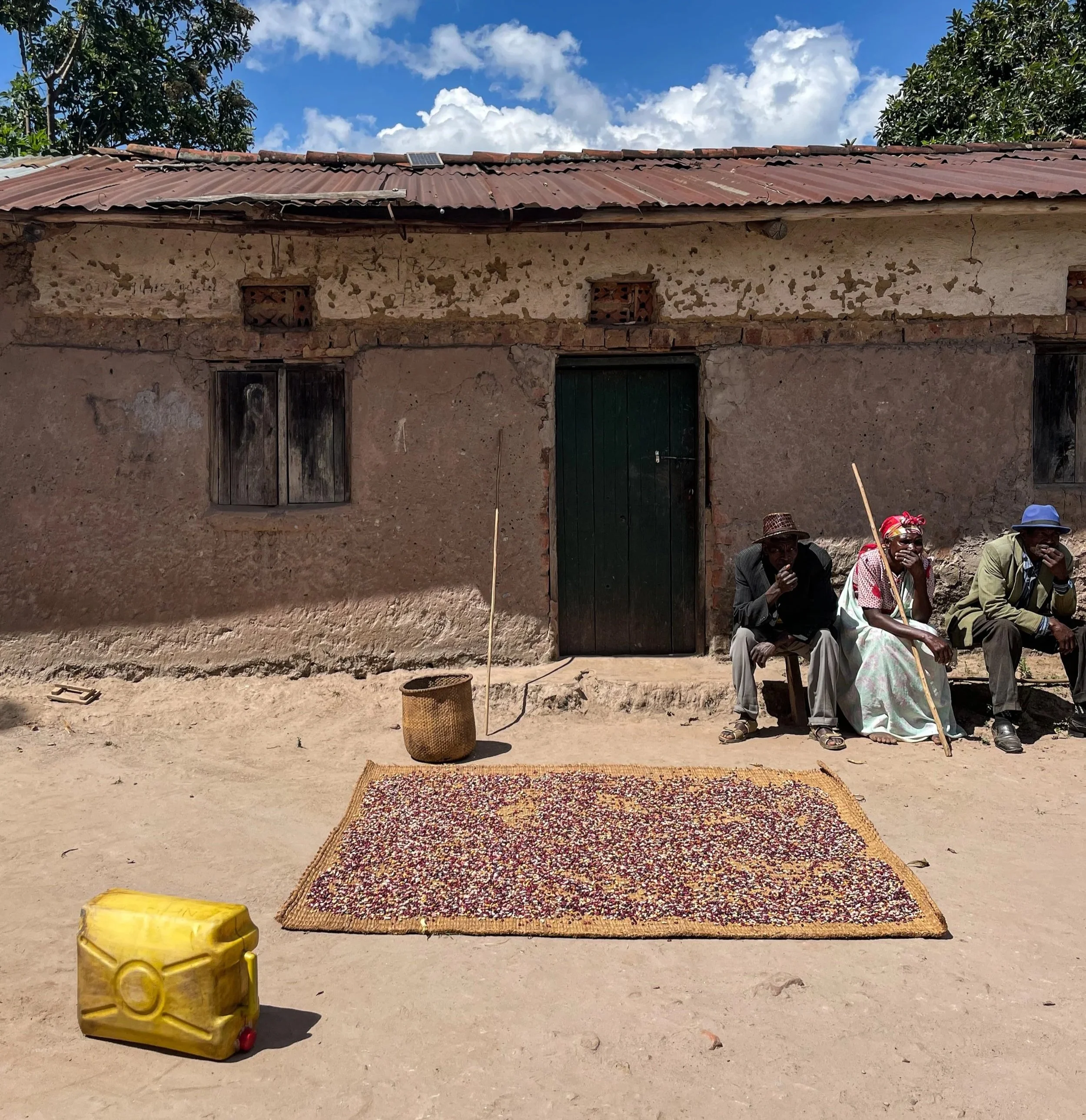 Three people sitting outside a weathered, mud-brick house with a rusty metal roof. A tarp is laid on the ground in front of them, with a yellow jerrycan and a woven basket nearby. The sky is partly cloudy, and trees are visible behind the house.