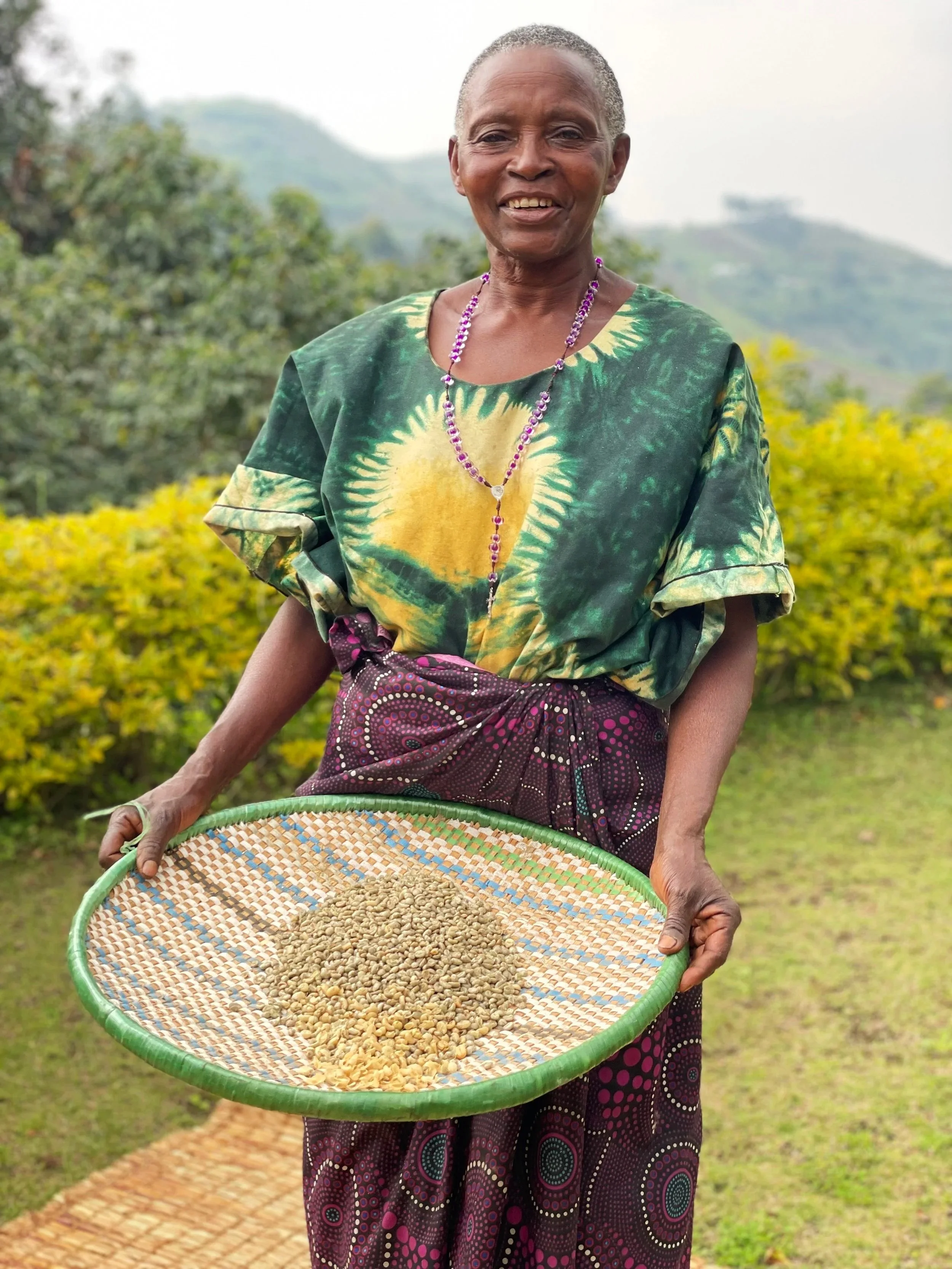 A woman smiling outdoors, holding a woven tray with various grains, dressed in vibrant traditional clothing with a green and yellow patterned top and purple patterned skirt, wearing a purple beaded necklace.