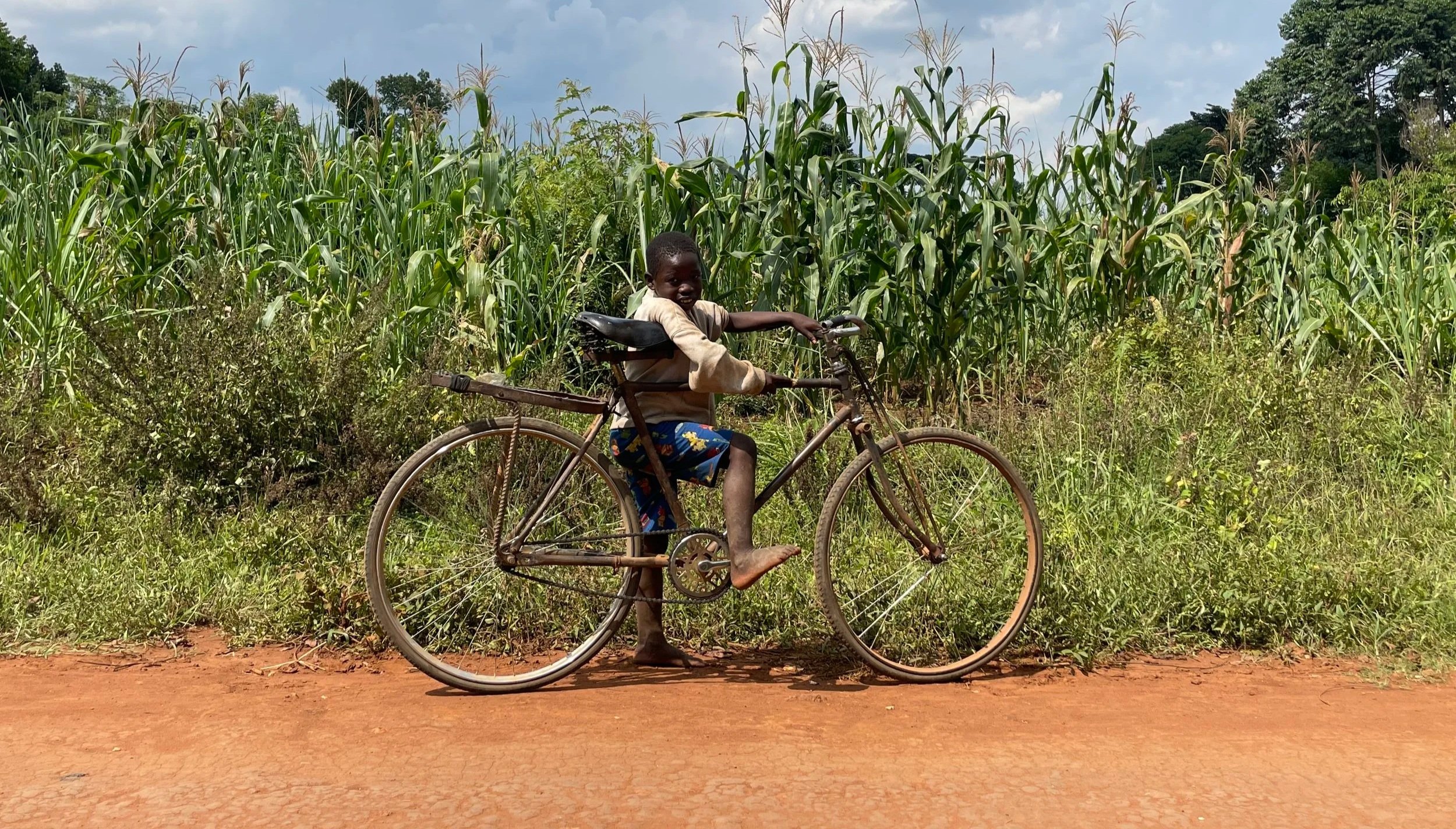 A young boy standing next to a bicycle on a dirt road with tall green crops and trees in the background under a partly cloudy sky.