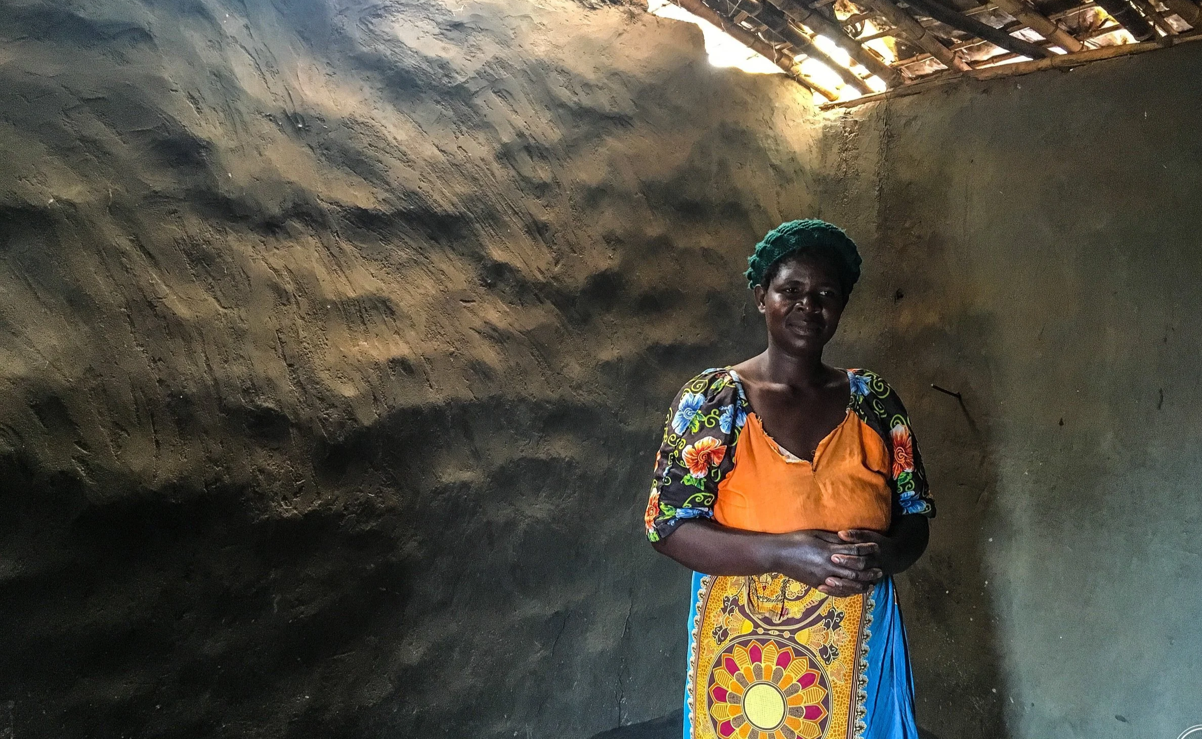 A woman standing inside a modest, unfinished room with dark, textured walls and a thatched roof partially open to the sky, wearing colorful traditional clothing.