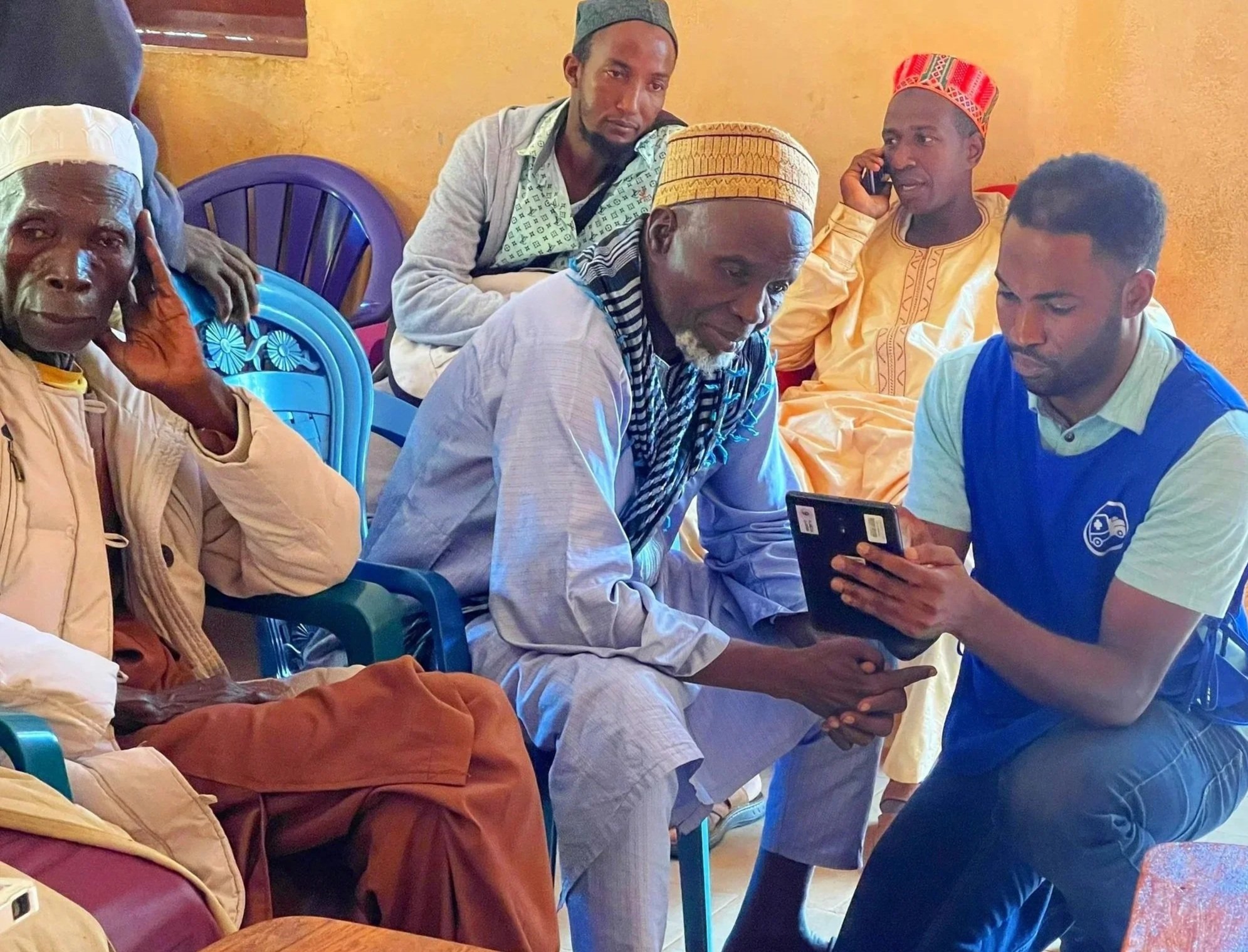 Group of men seated and engaging with a smartphone, in a room with orange walls. Some men wear traditional clothing and hats, while others wear casual Western attire.