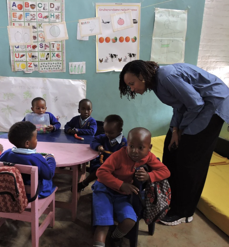 A teacher interacts with five young children in a classroom. The children are seated around a pink table and a small chair. The classroom has educational posters on the wall, including the alphabet, pictures of vegetables and animals, and some drawings. The children wear school uniforms, and the scene captures a lively learning environment.