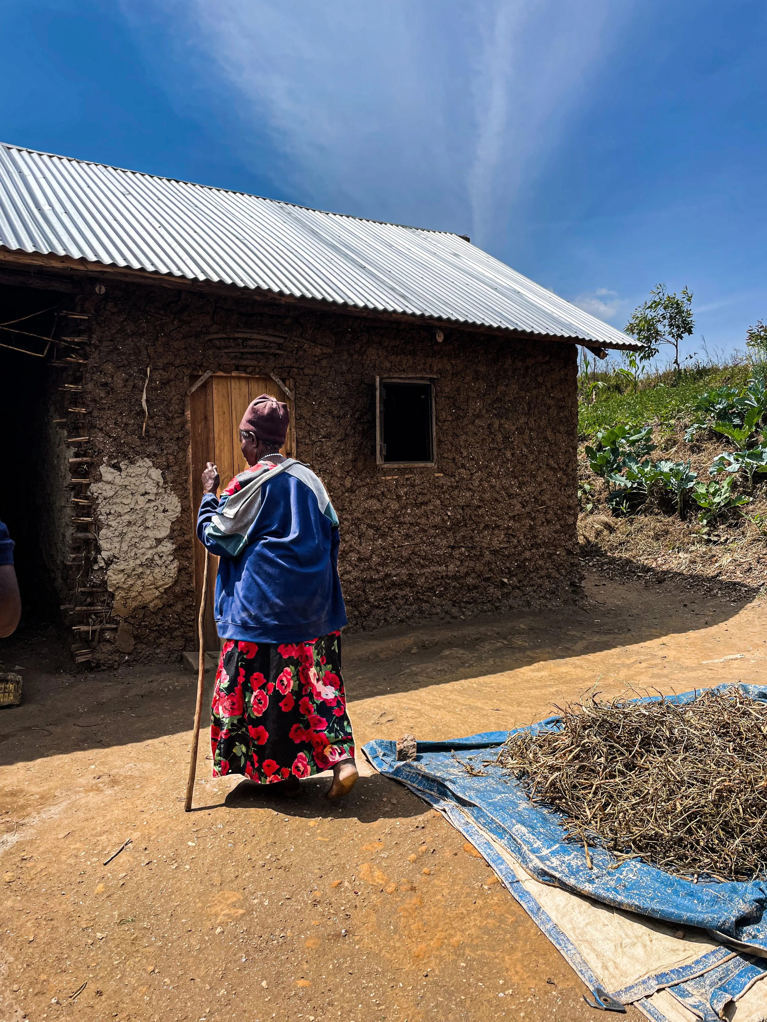 An older woman in a floral skirt and purple beanie standing outside a mud house with a metal roof, and a blue tarp bed of dried plant material nearby, under a clear blue sky.