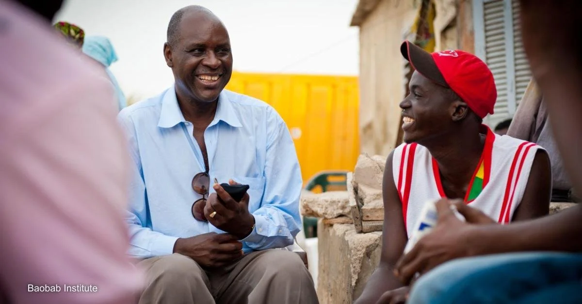 Two men are smiling and talking outdoors, with other people partially visible around them, in an informal setting with a yellow wall and a building in the background.