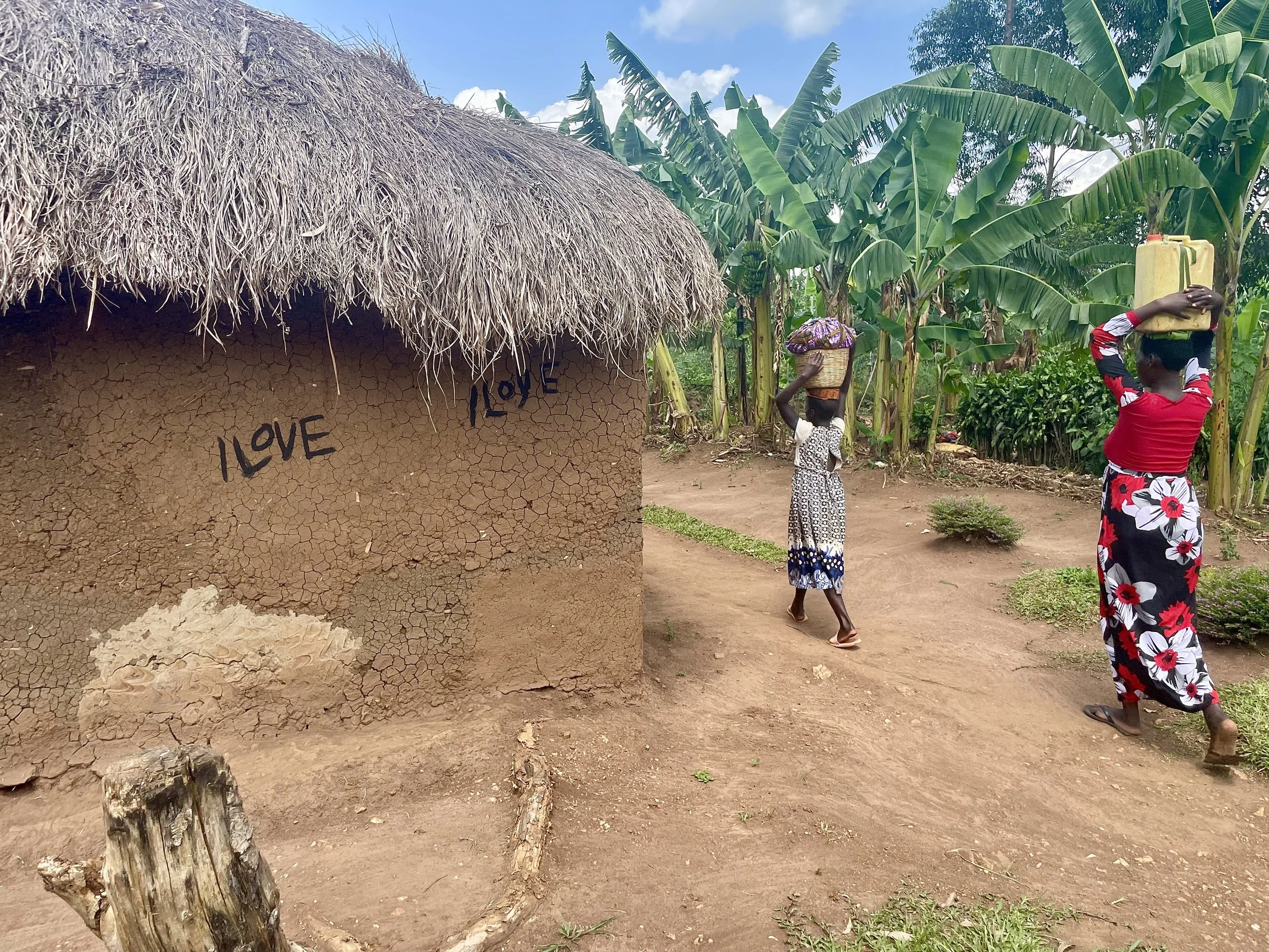A dirt path next to a traditional mud house with a thatched roof. The house has the word "LOVE" painted on the wall. Two women are carrying containers on their heads, walking along the path. One woman is dressed in a red top and floral skirt, the other in a white and black dress. There is lush green vegetation and banana plants in the background.