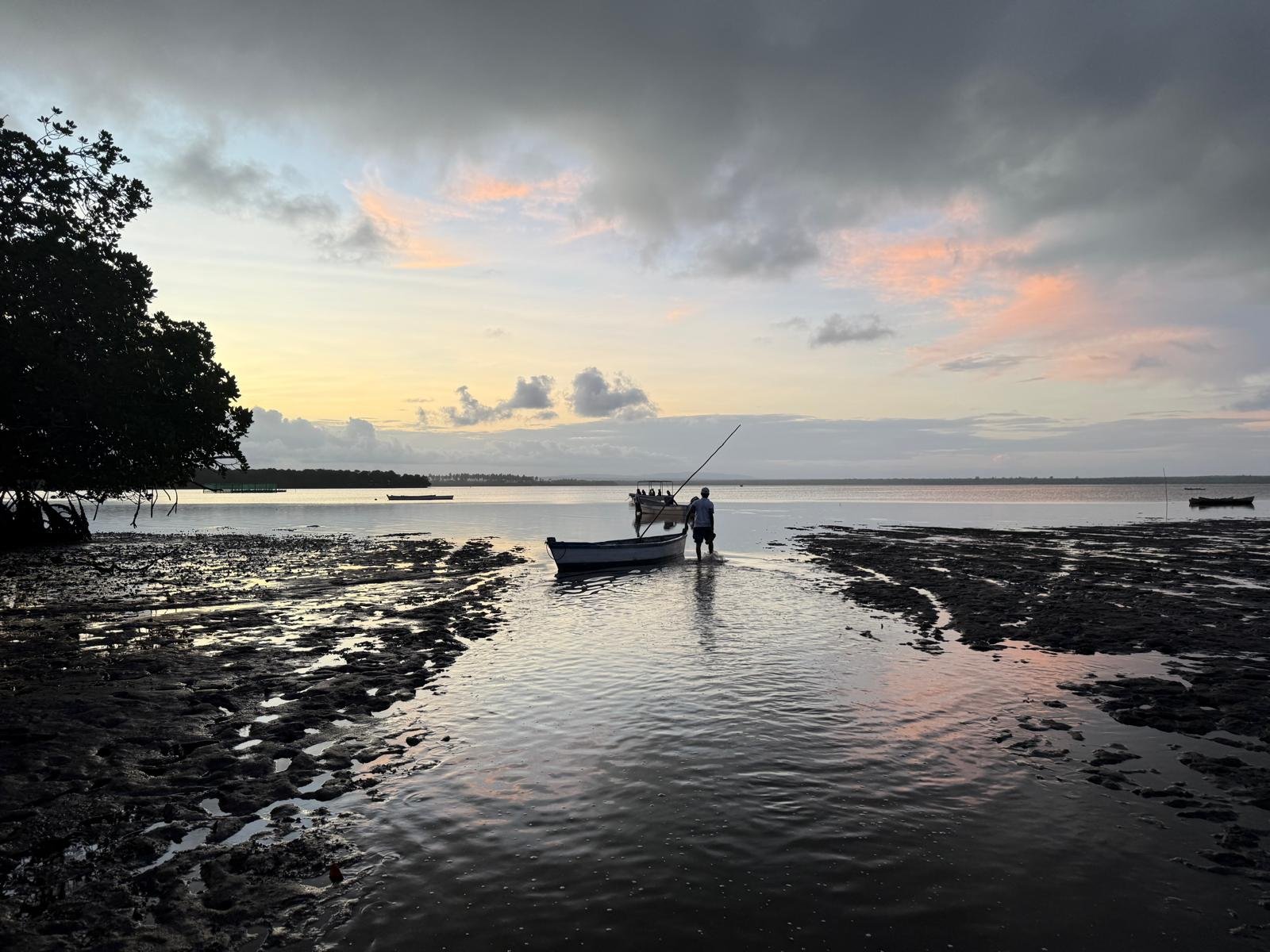 A person standing in shallow water near a small boat on a calm river at sunset, with boats in the distance and a cloudy sky with pink and orange hues.