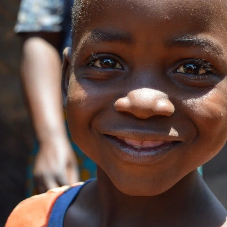 Close-up of a smiling young boy with dark skin and short hair, outdoors on a sunny day.