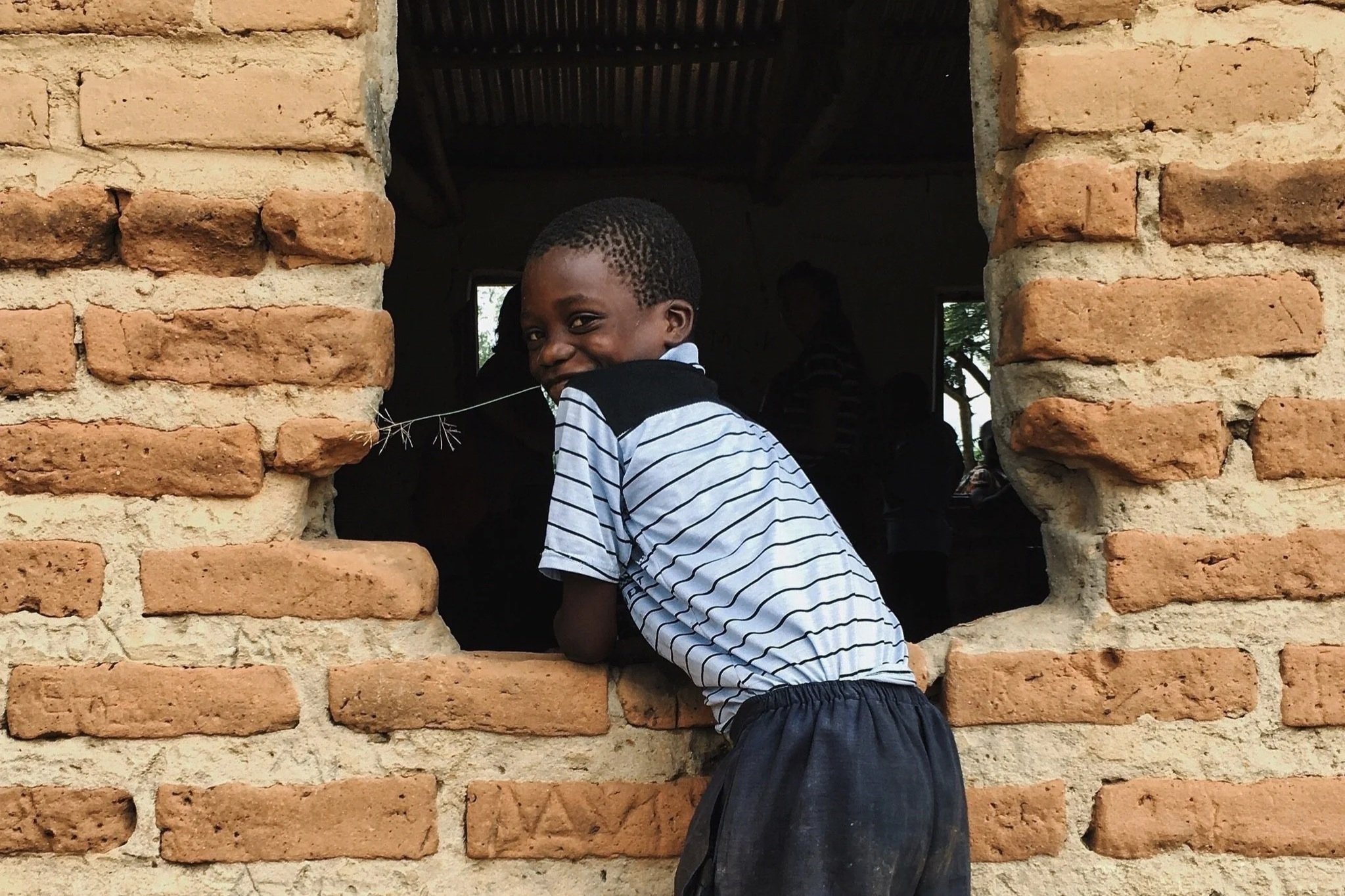 A smiling young boy wearing a striped shirt and shorts leaning on a brick window ledge, looking into the camera, with a dark interior and other children visible in the background.