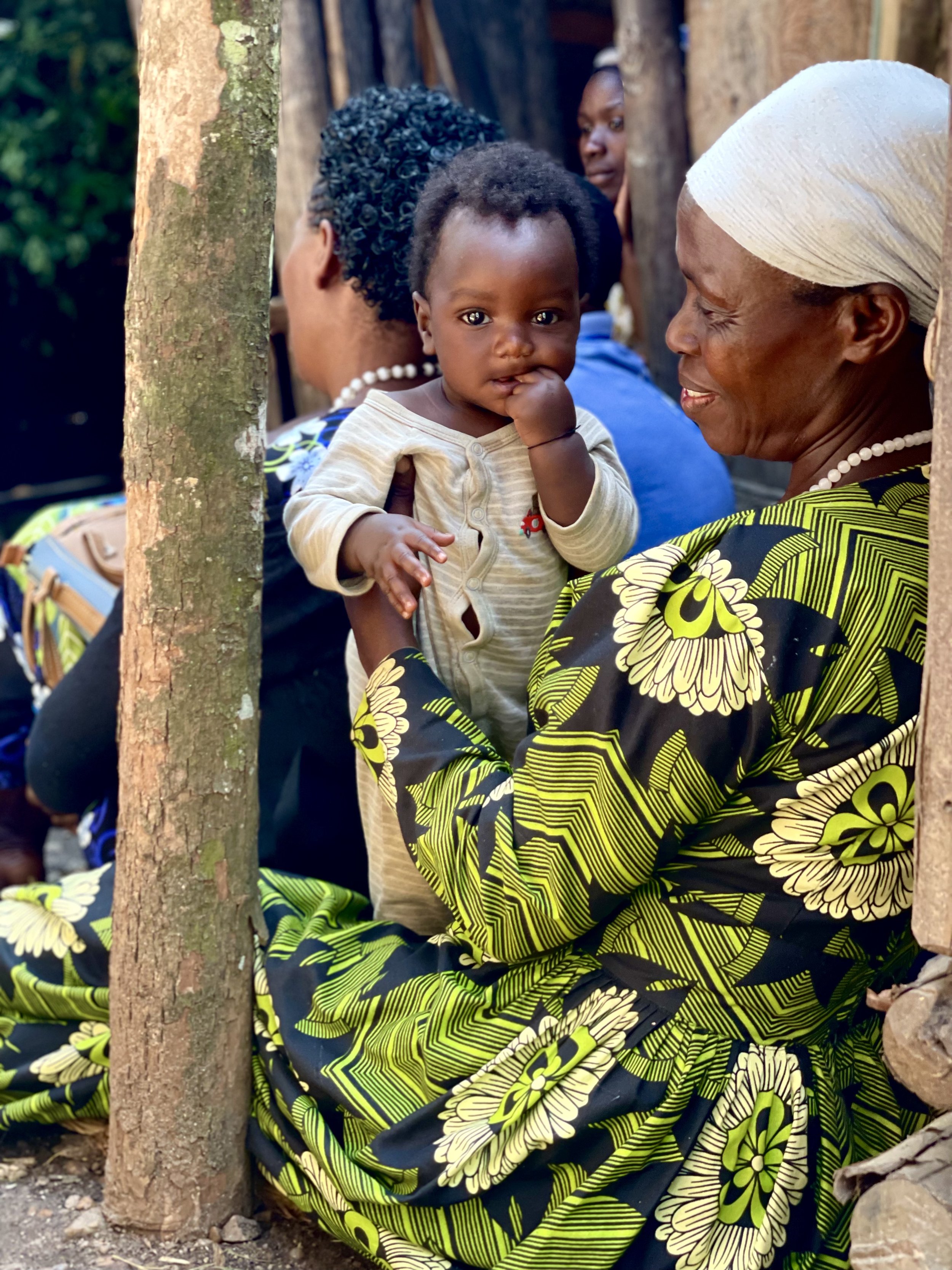 A woman with a white headscarf and a black and yellow floral patterned dress holds a young child with dark curly hair and a striped beige shirt. The woman is smiling, and the child is resting his hand on his mouth, looking at the camera. They are sitting outdoors near a wooden post, surrounded by other women.