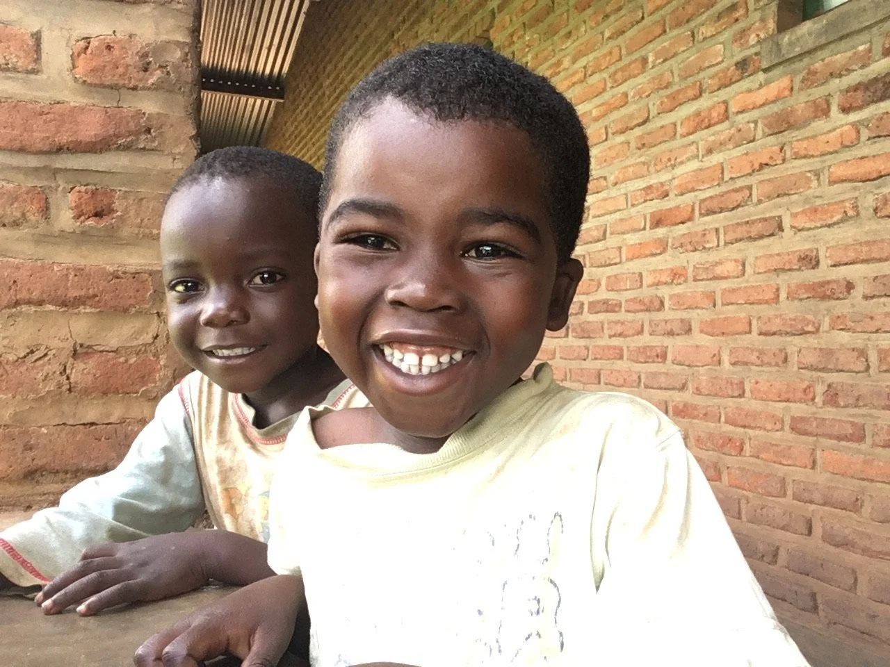 Two smiling young boys sitting at a table outdoors with a brick wall background.