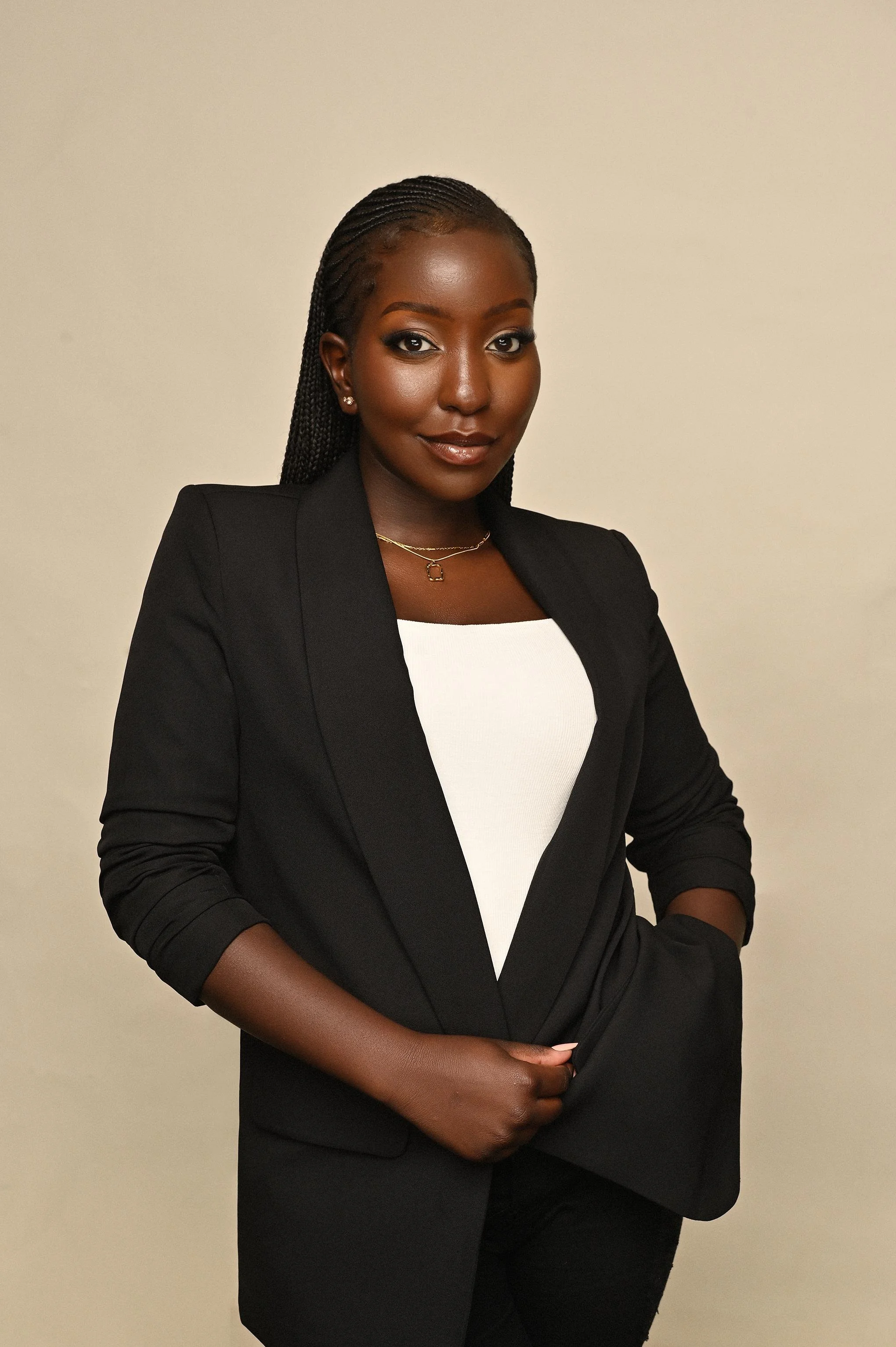 A woman with braided hair, wearing a black blazer over a white top, standing with her hand in her pocket against a plain beige background.