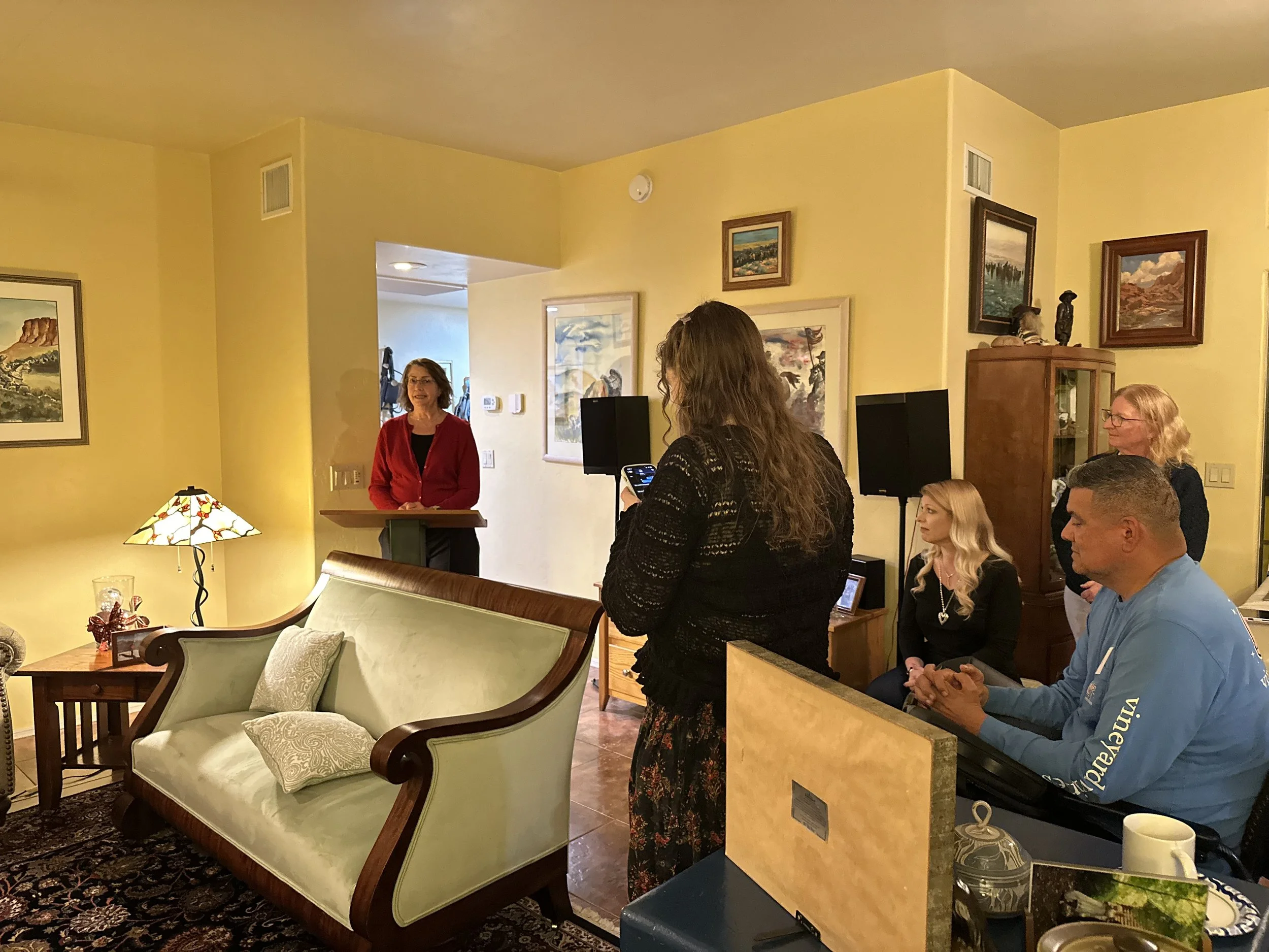 A woman in a red cardigan stands at a small wooden podium in a living room, speaking to an audience. The audience includes a woman with long brown hair holding a phone, another woman with blonde hair sitting on a chair, a man in a blue shirt sitting 