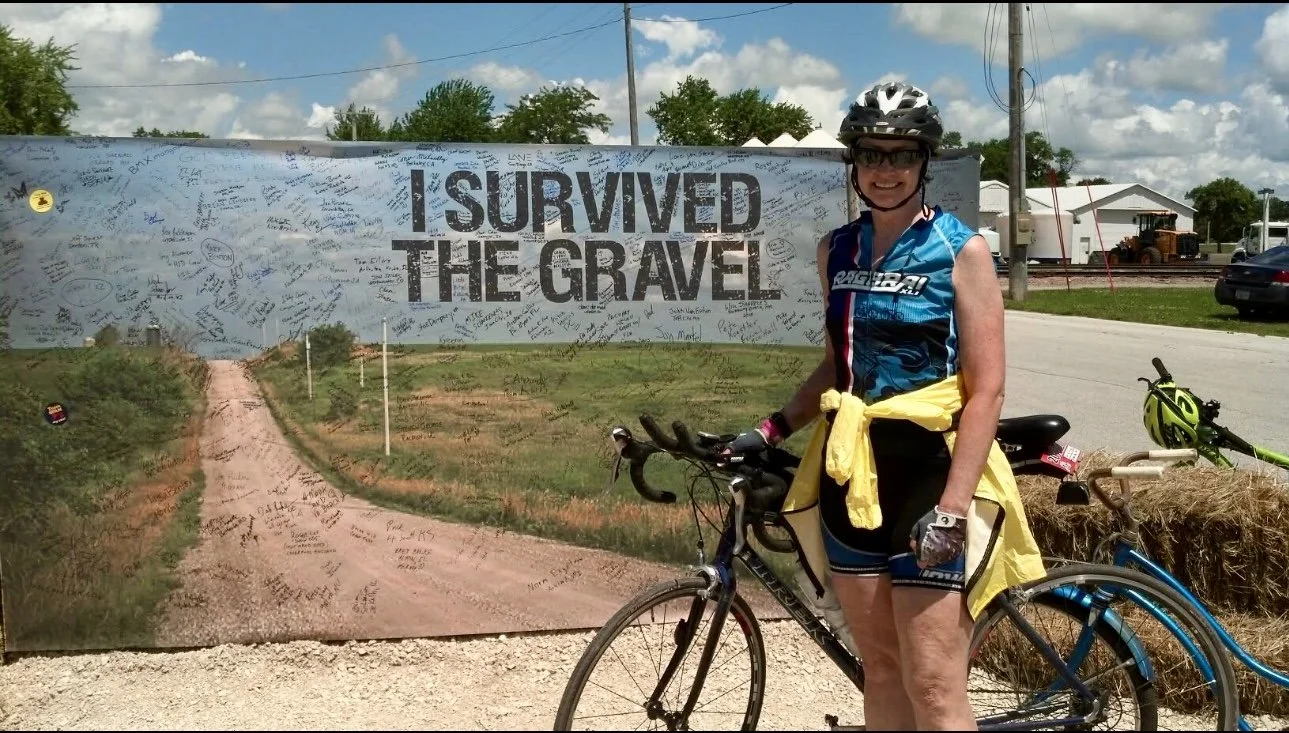 Julie in cycling gear standing next to her bicycle in front of a large sign that reads 'I SURVIVED THE GRAVEL' at a gravel cycling event, with barn and tractor in the background.