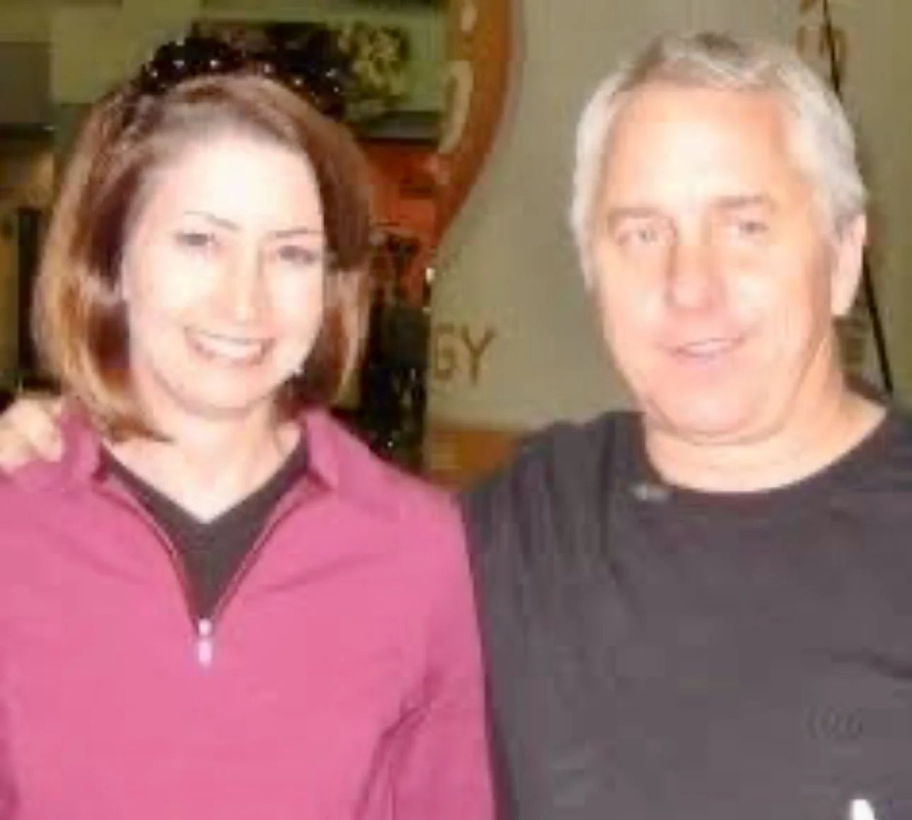 Julie wearing a pink zip-up jacket and a man with gray hair wearing a black shirt, smiling and standing close together indoors.