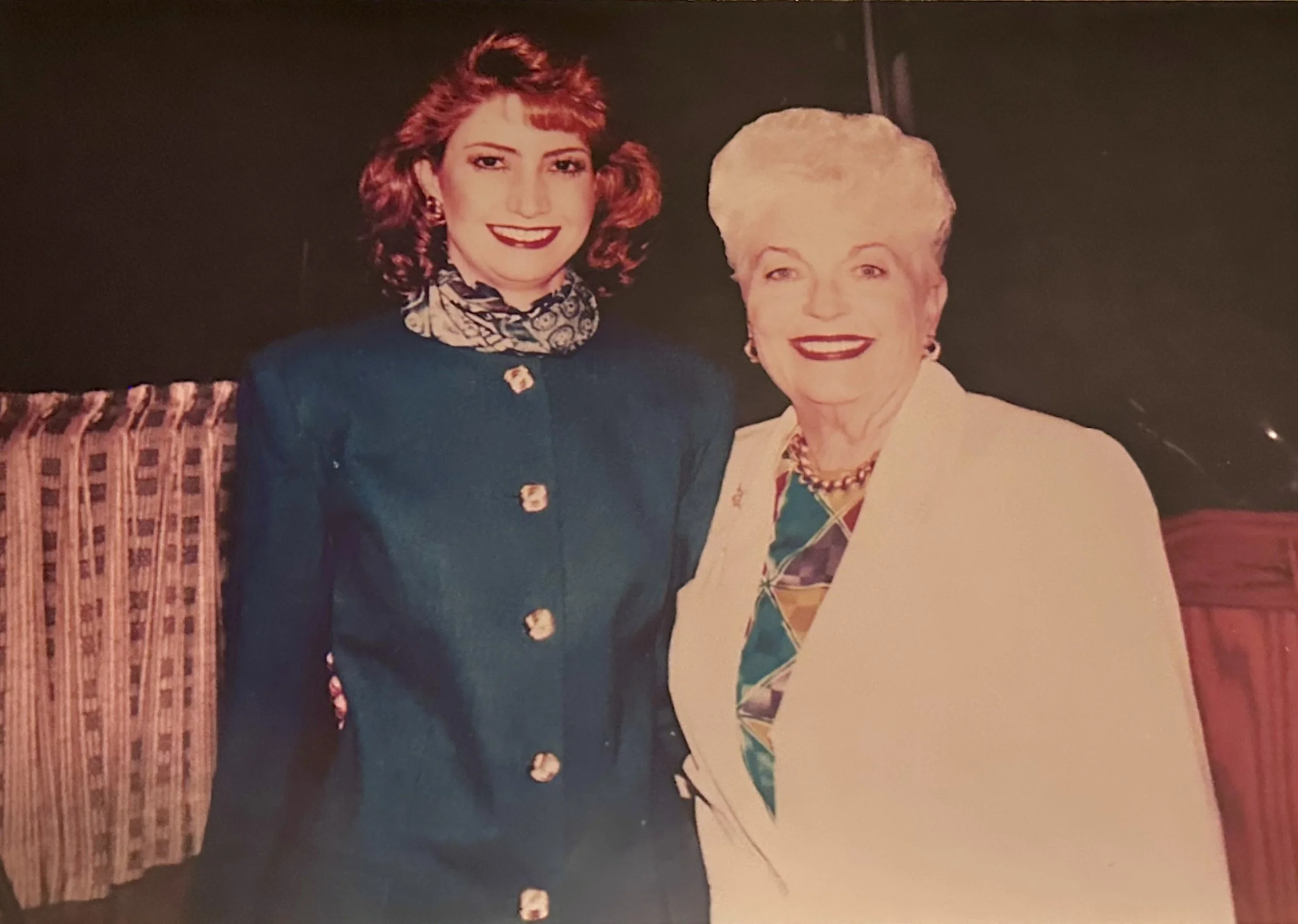 Two women are posing together indoors, smiling at the camera. The woman on the left has red hair and is wearing a dark navy blazer and a patterned scarf. The woman on the right has light-colored hair and is dressed in a white blazer with a colorful p