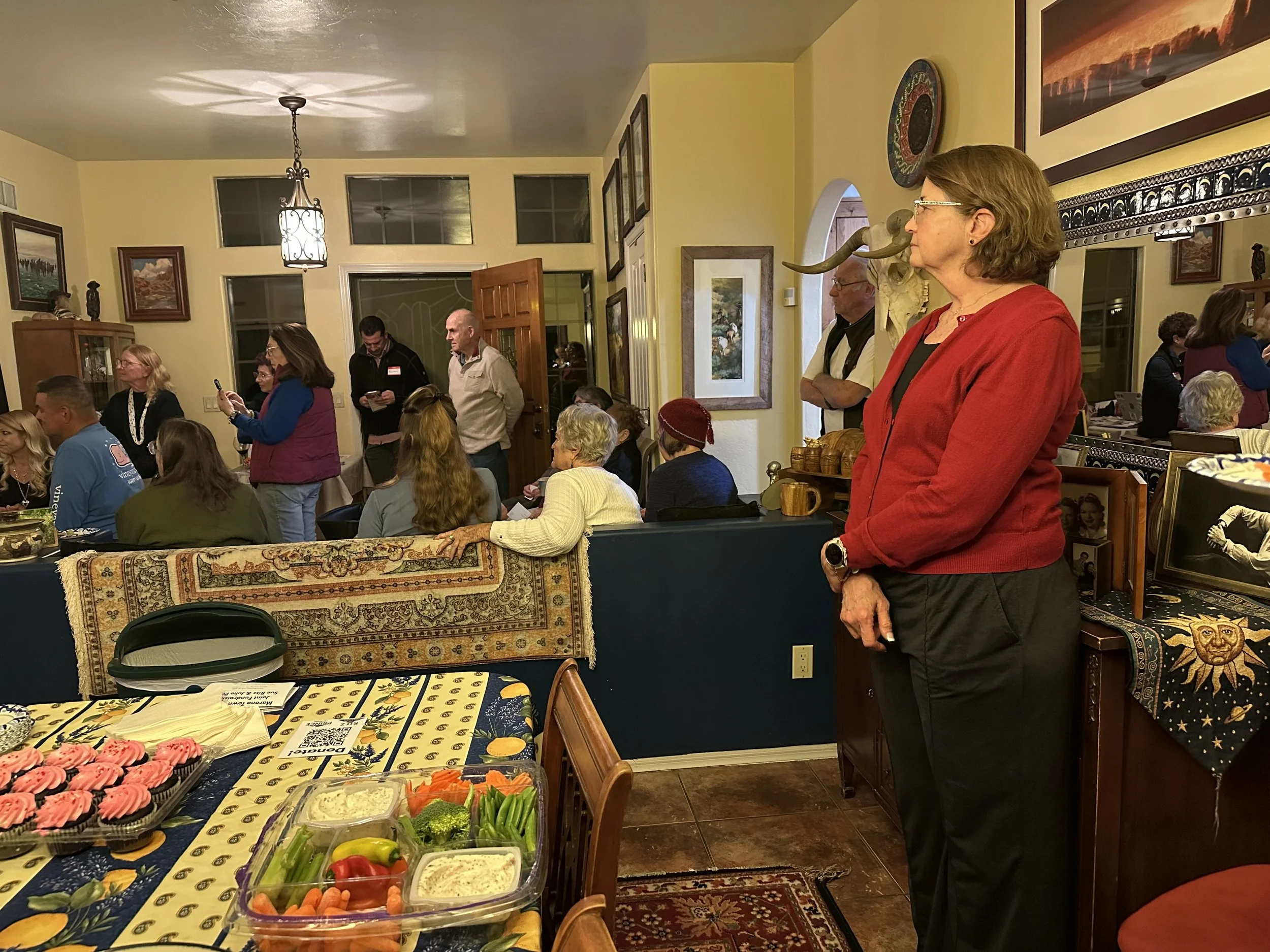 A group of people gathered in a warmly lit living room, some seated at a table with food, others standing and listening. A woman in a red jacket stands to the right, facing the left. The room has framed art and a large animal skull on the wall, with 