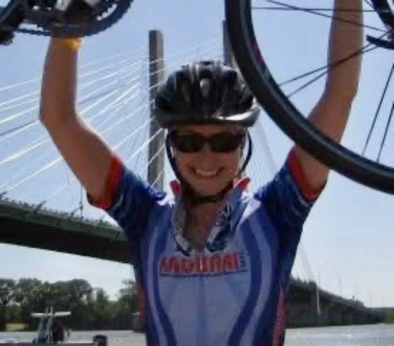 Julie wearing sunglasses, helmet, and cycling jersey, holding up a bicycle wheel with arms raised, smiling, outdoors under a bridge.