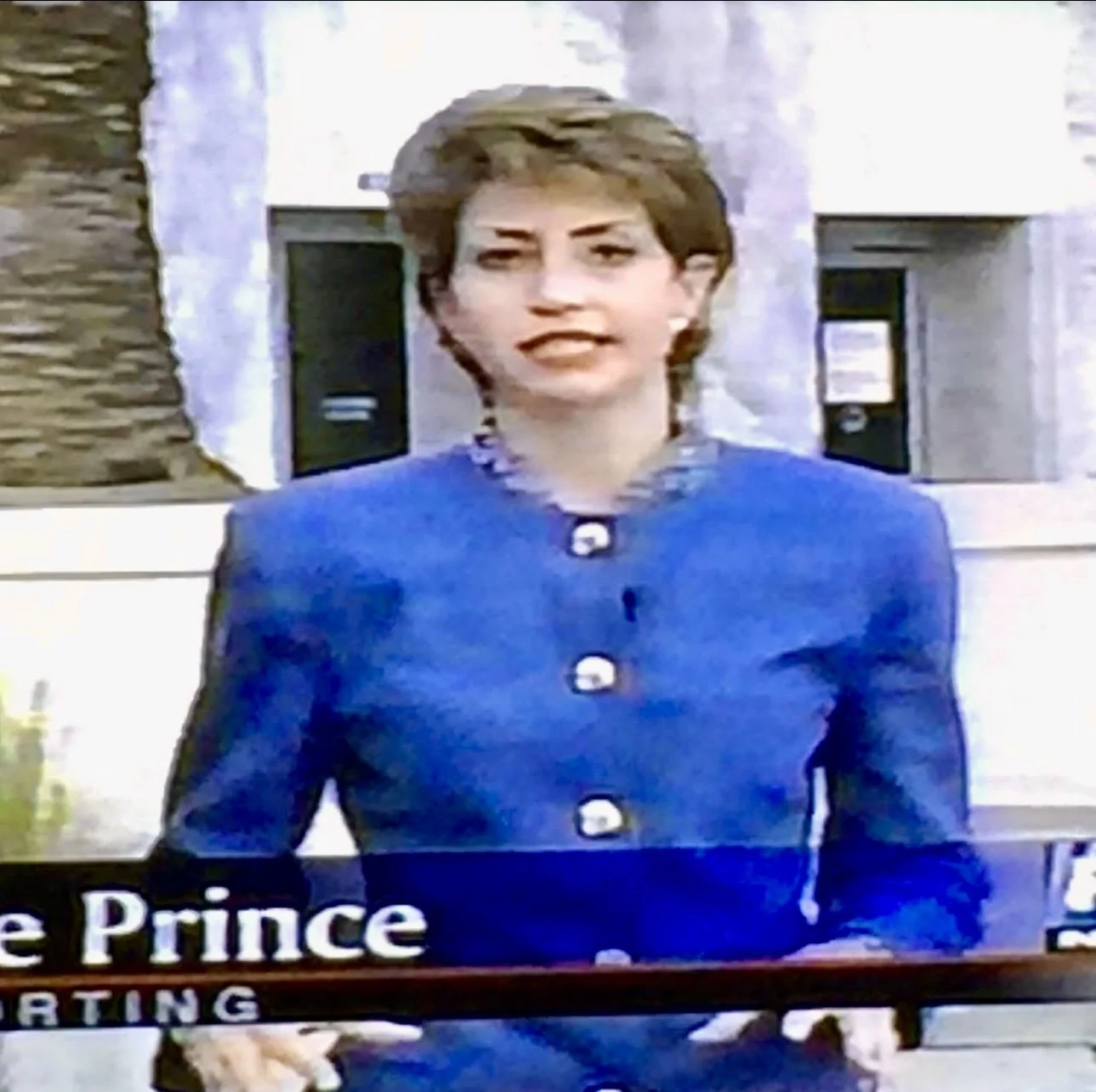 Julie wearing a bright blue blazer with large white buttons, standing outdoors in front of a building with white walls and small trees, possibly on a TV set or during a news broadcast.