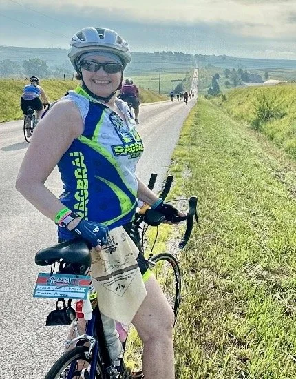 Julie in cycling gear, wearing a helmet and sunglasses, standing with her bicycle on a rural road surrounded by green fields, with other cyclists in the background under a cloudy sky.