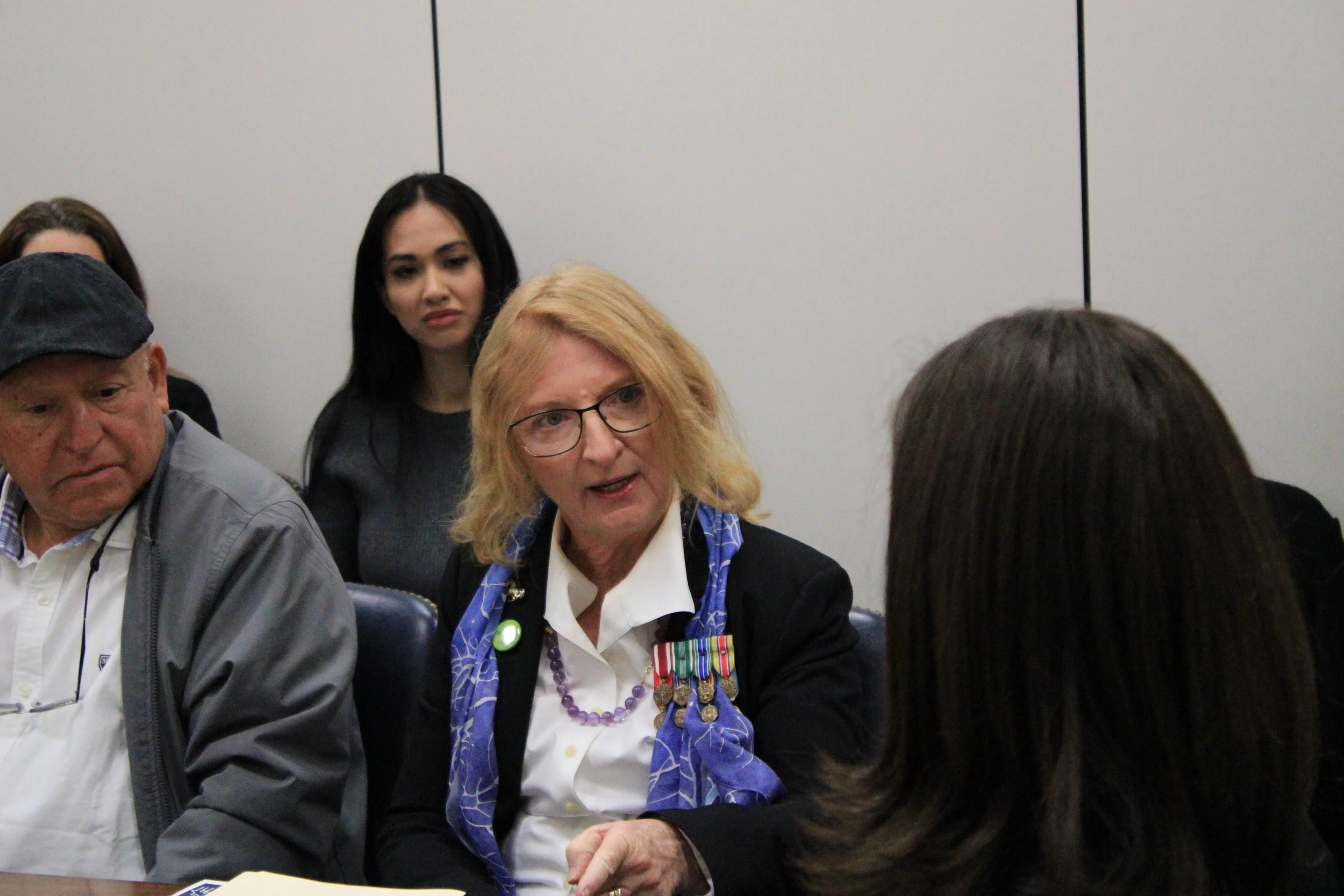 Sue wearing medals on her jacket, a man with a cap, and women with dark hair, in an indoor setting with a white wall background.