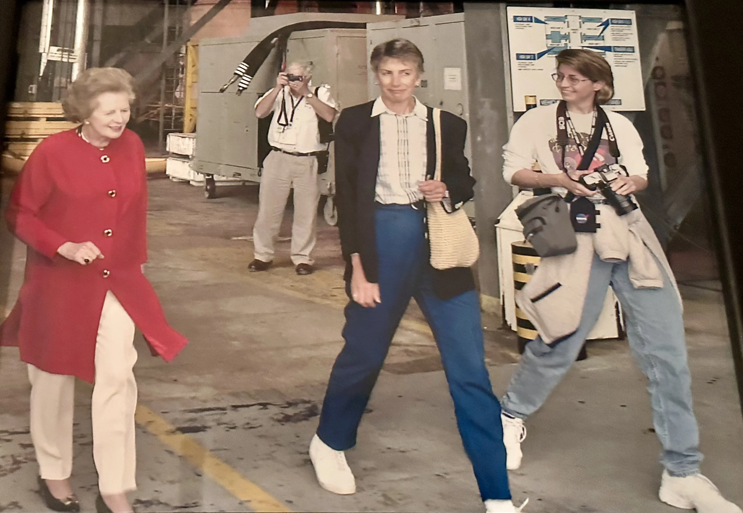 Three women in an industrial or warehouse setting, one in a red coat, one in a suit with blue pants, and the third with a camera strap around her neck and holding a camera, with a woman taking a photo in the background.