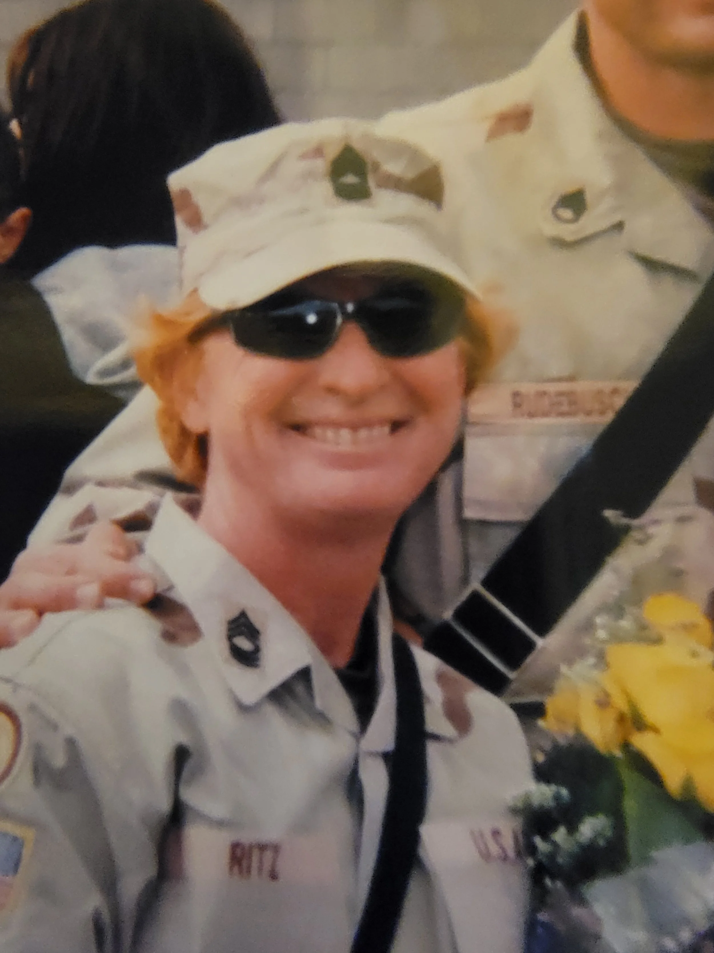 Sue in military uniform, wearing sunglasses and a hat, smiling, holding a bouquet of yellow flowers.