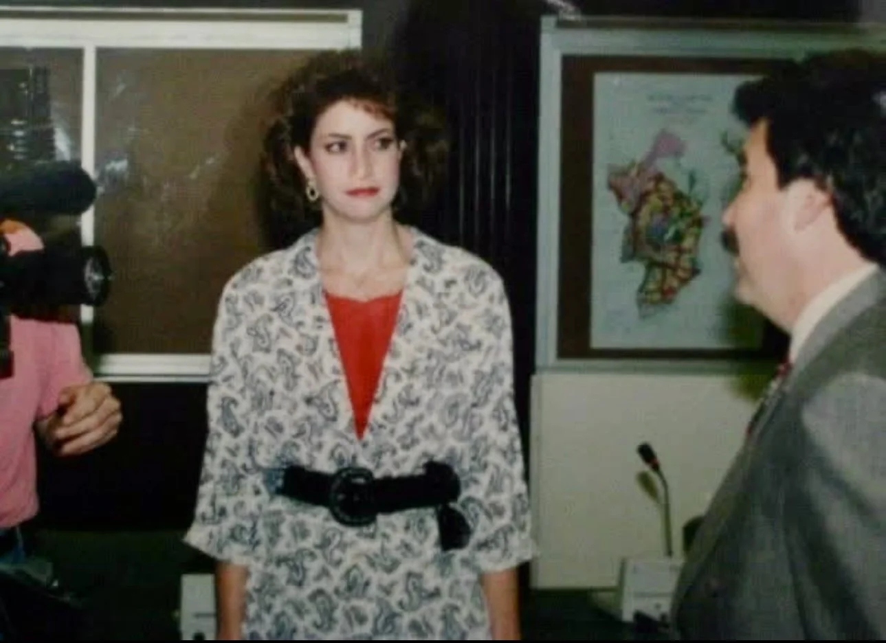 Julie with earrings wearing a white paisley-patterned blazer over a red top, standing and looking to her right, in a room with a map of Ireland in the background. A man with dark hair in a gray suit is facing her on her left.