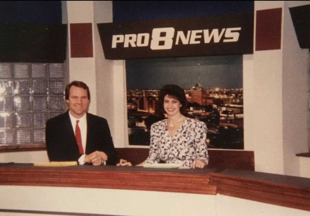 Two news anchors, a man in a black suit and red tie and a woman in a patterned blouse, sitting behind a news desk with a cityscape background and a 'Pro 8 News' sign above them.