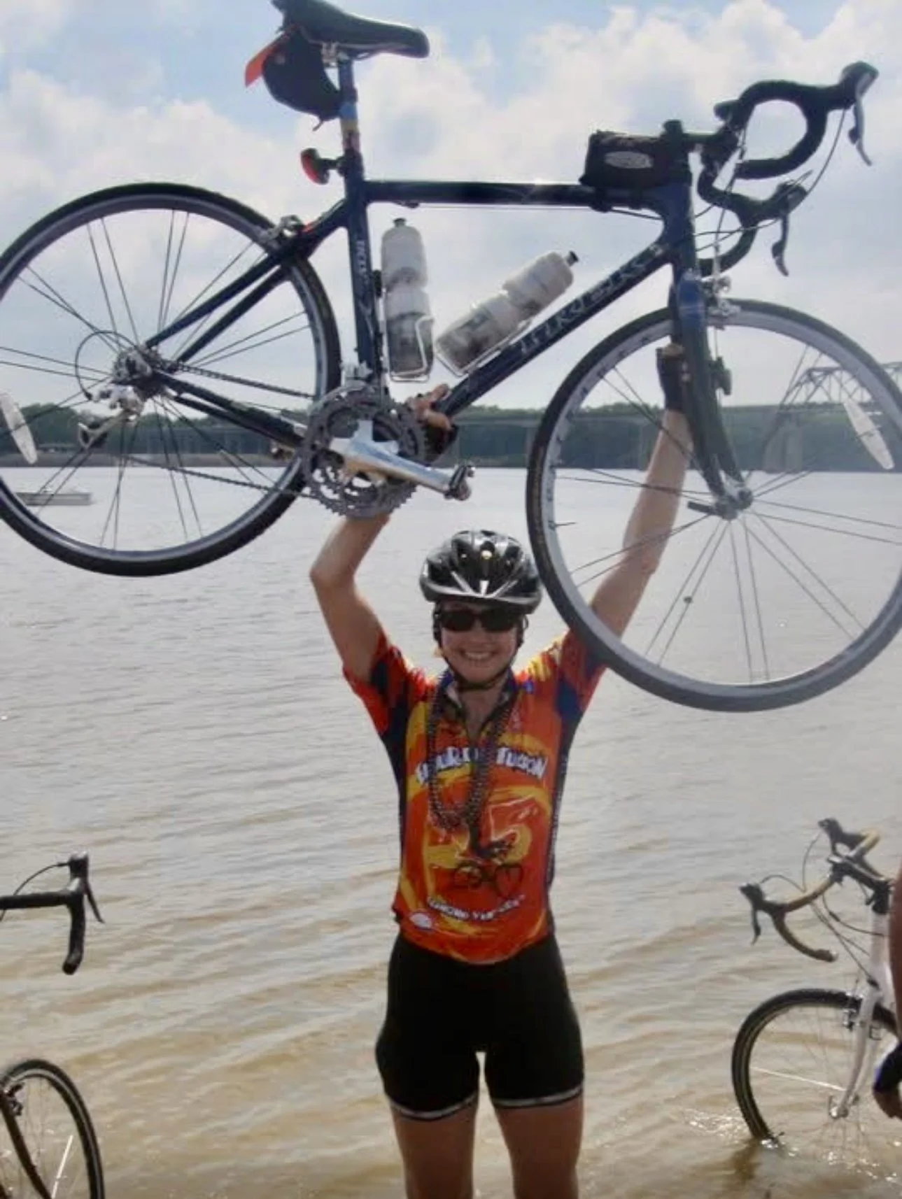 Person in cycling gear and helmet holding a bicycle above their head by the water with other bicycles nearby.