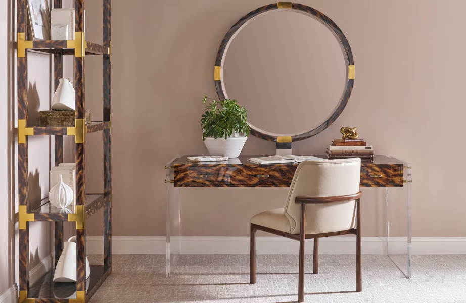 A home office setup with a marble and wood desk, a round mirror above, a beige chair, a bookshelf with decorative items, and a potted plant.