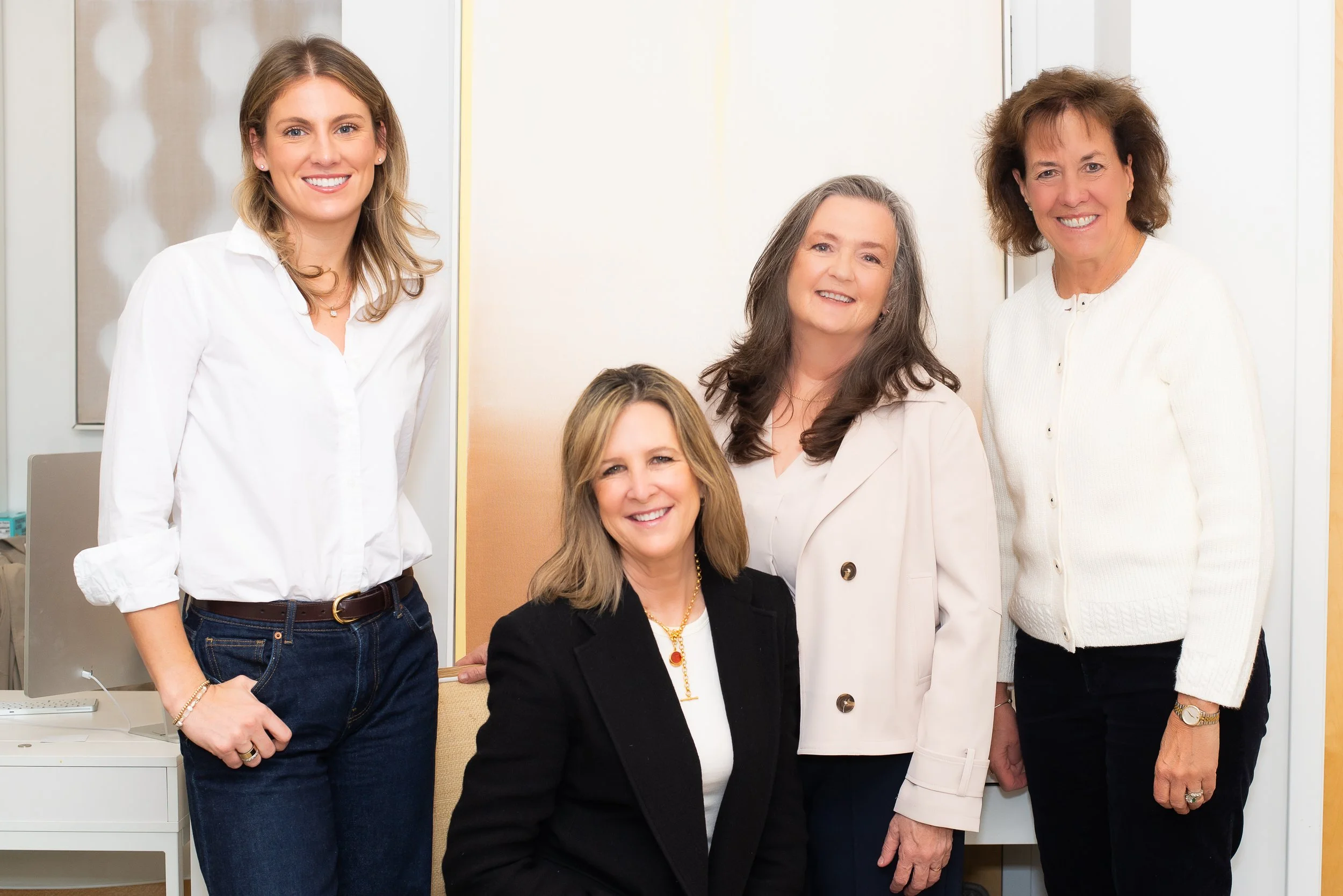 Five women smiling and posing together in an office setting, some standing and one sitting, dressed in business casual attire.